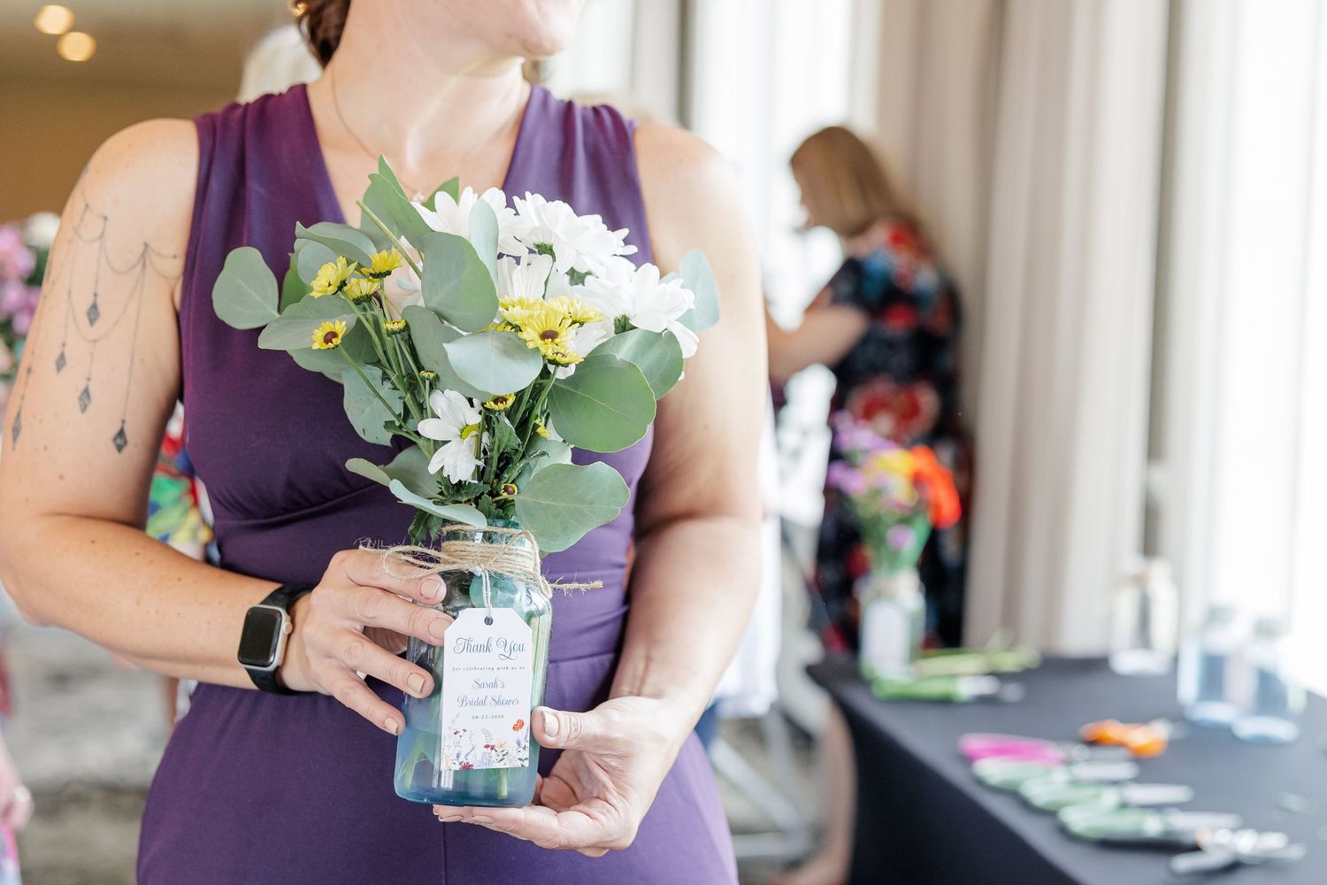 Woman in purple dress holding a mason jar vase with white and yellow flowers at an indoor event.