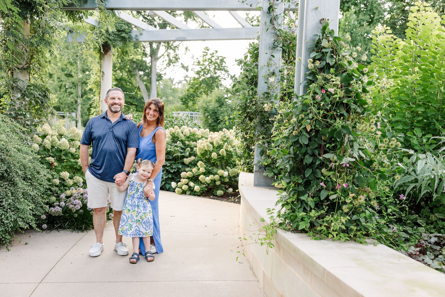 Family of three, parents and young daughter, smile together under a pergola surrounded by lush green gardens.
