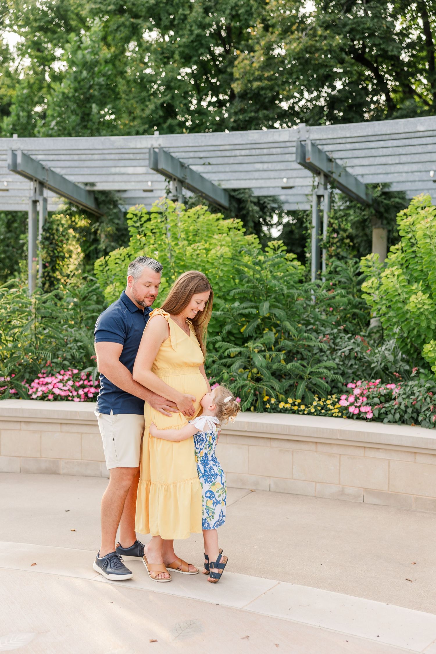 Pregnant couple in a garden holding their toddler daughter between them as she touches moms baby bump.