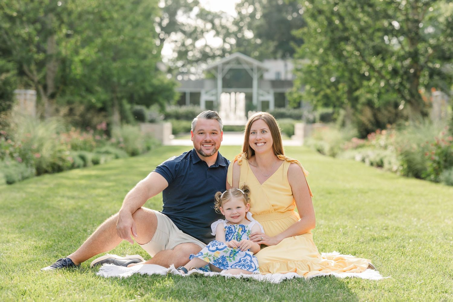 Happy family of three sitting on blanket in garden with gazebo backdrop on sunny day.