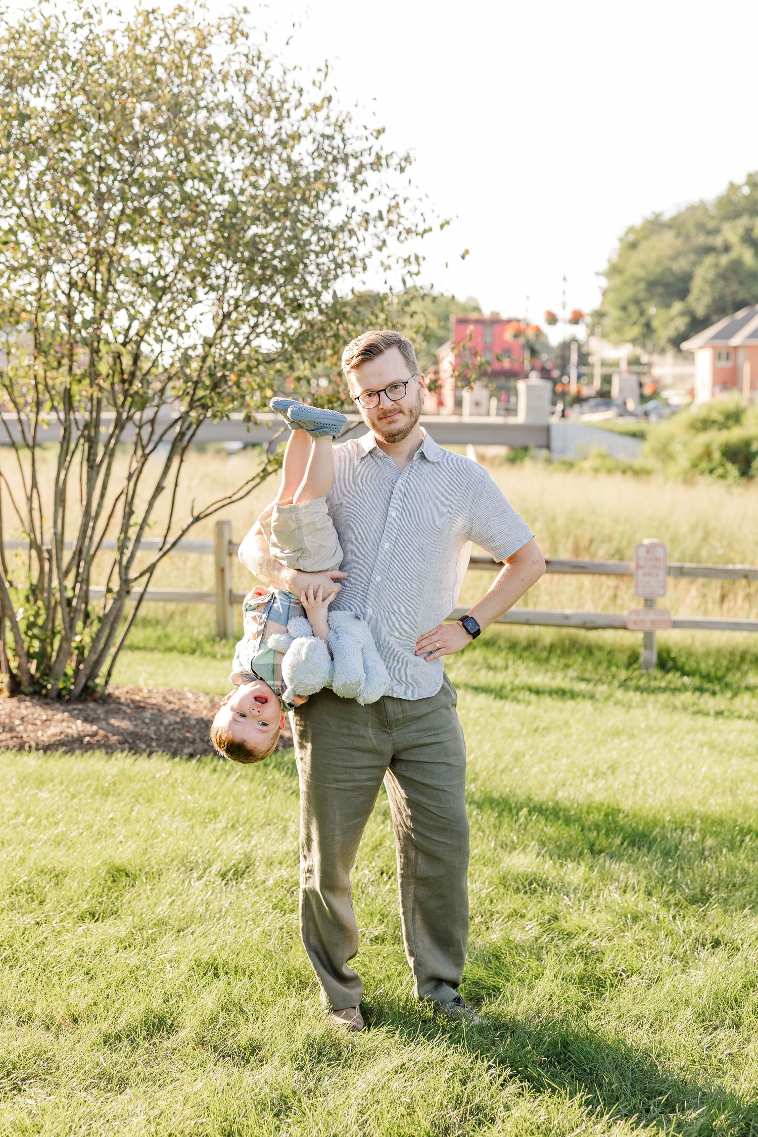 A playful father holds his young child upside down while laughing outdoors near a wooden fence and shrub.