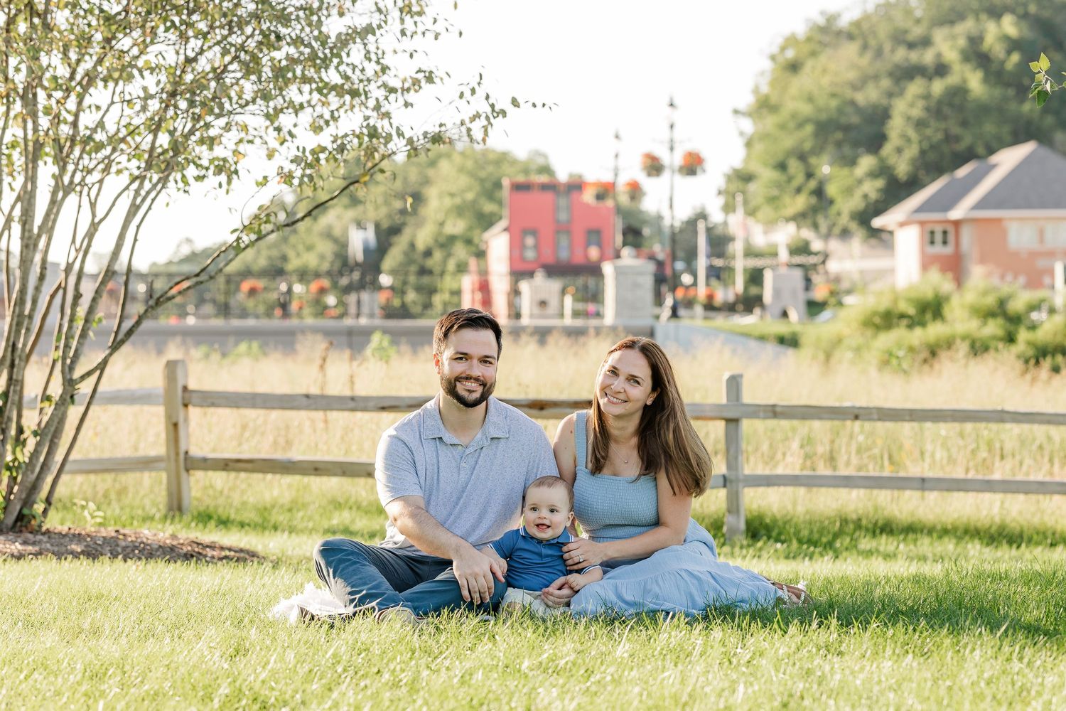 A young family with a baby sits on green grass in front of a wooden fence on a sunny day.