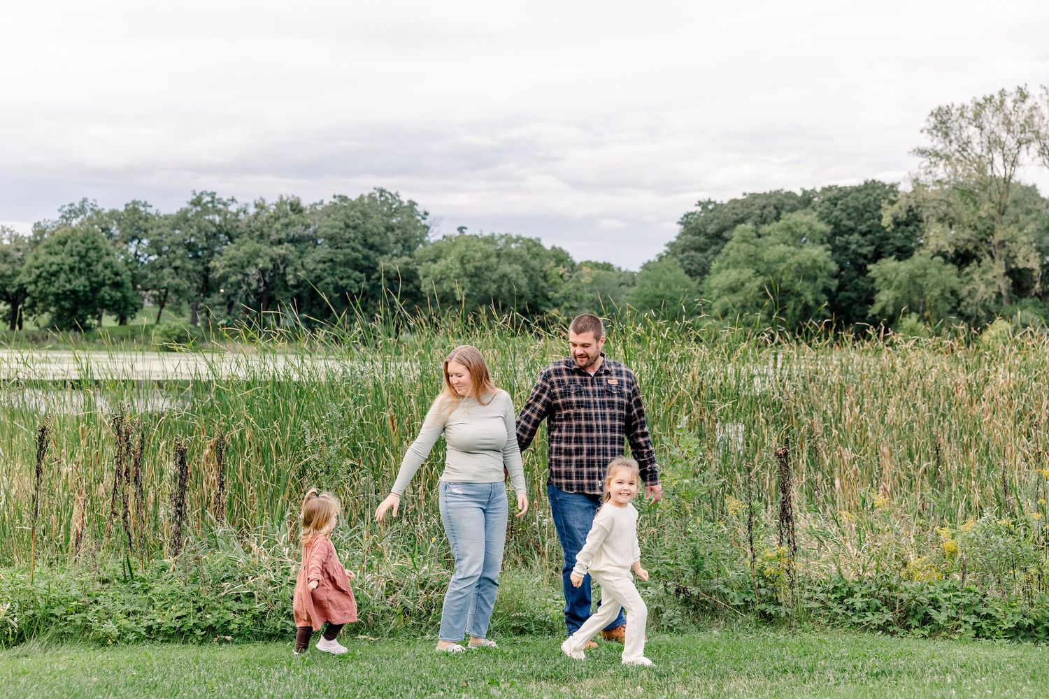 A family of four walks hand in hand through a lush green field beside tall reeds near a pond.