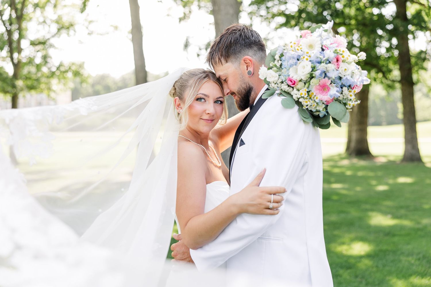 Bride and groom embrace outdoors with flowing veil, colorful bouquet, and lush green trees in background.