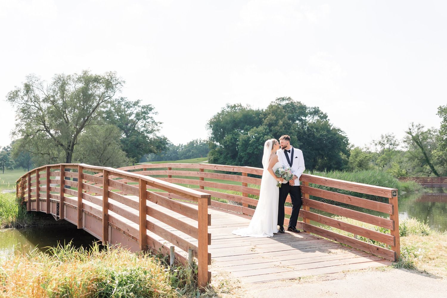 Bride and groom pose together on rustic wooden bridge surrounded by lush greenery.