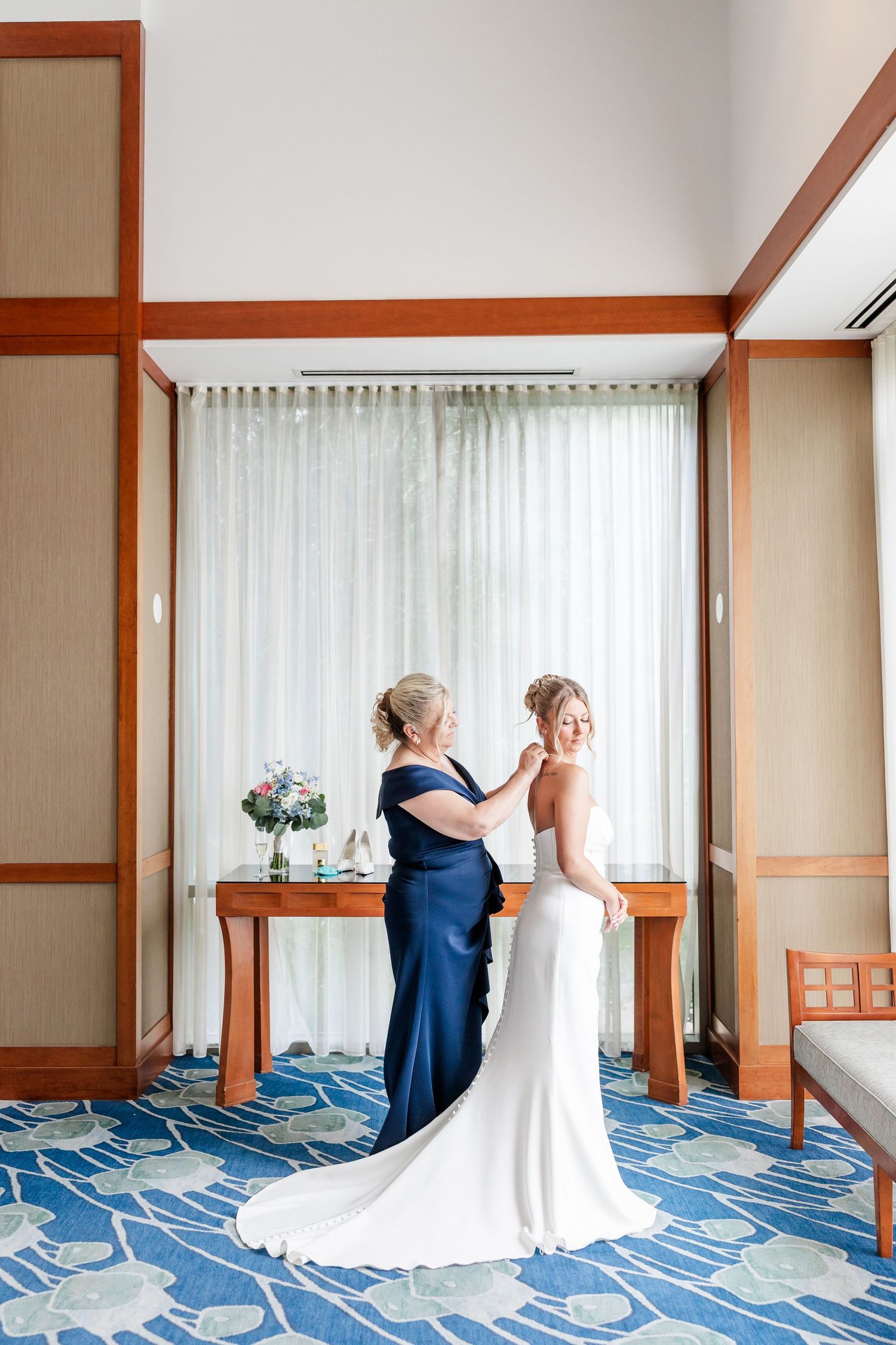 Mother helping bride with wedding dress in elegant hotel room with blue carpet and sheer curtains.