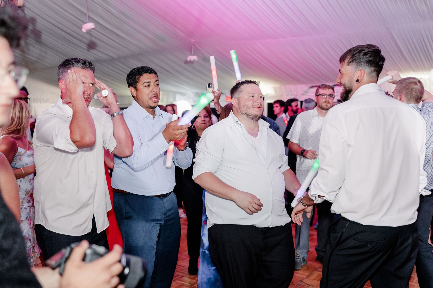Groomsmen and guests dancing with glow sticks at lively outdoor wedding reception under tent.