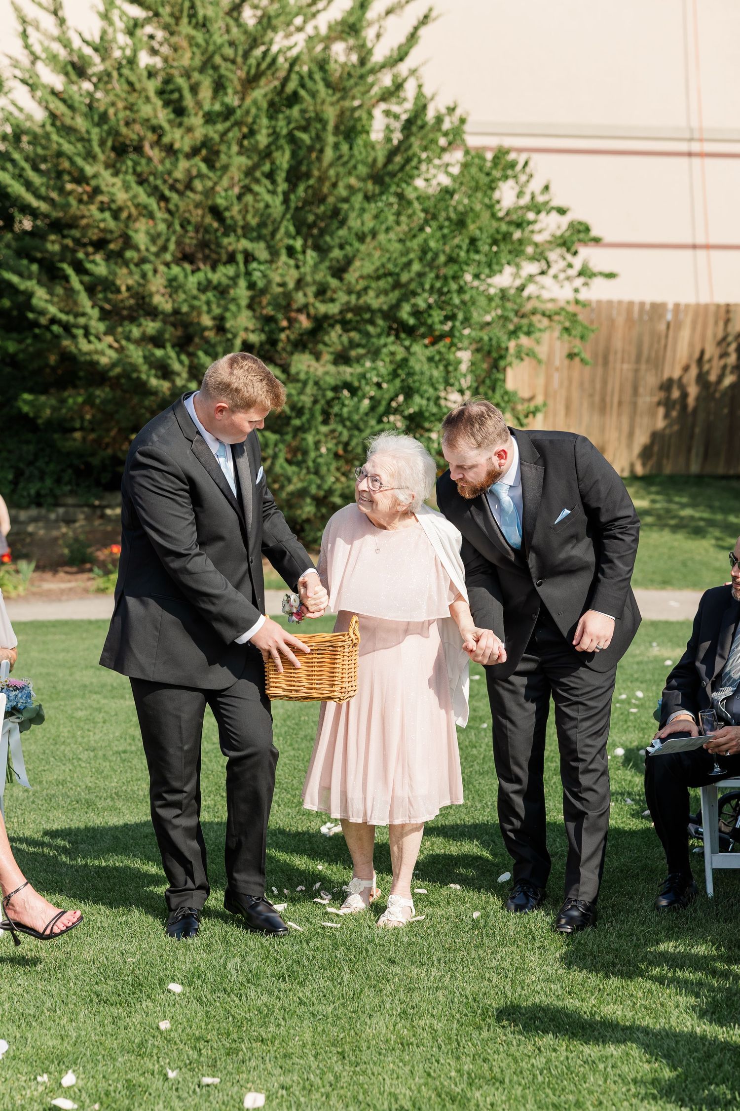 Elderly flower girl escorted by two groomsmen in black suits down outdoor wedding ceremony aisle.