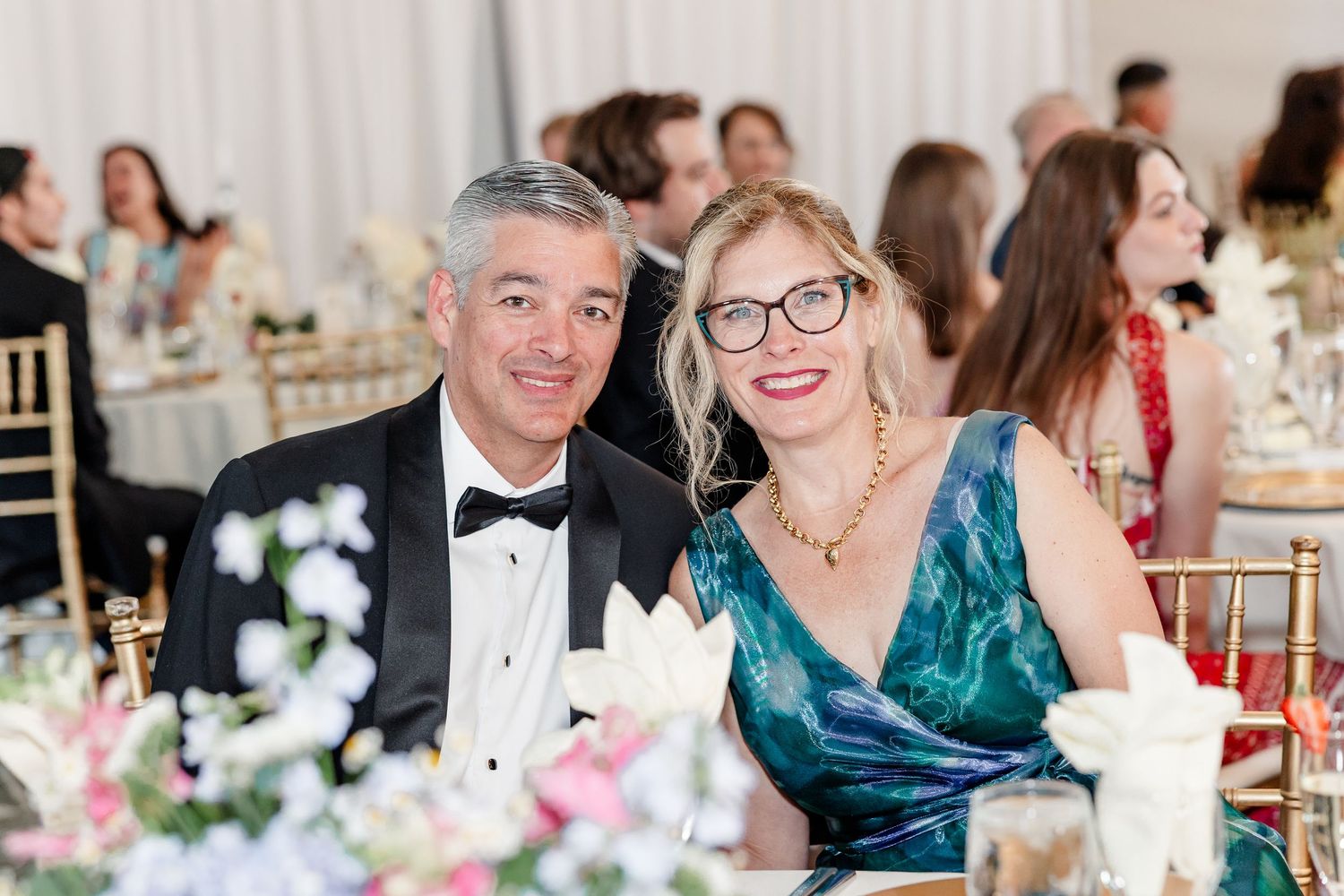 Smiling couple in formal attire seated at elegantly decorated wedding reception dinner table.