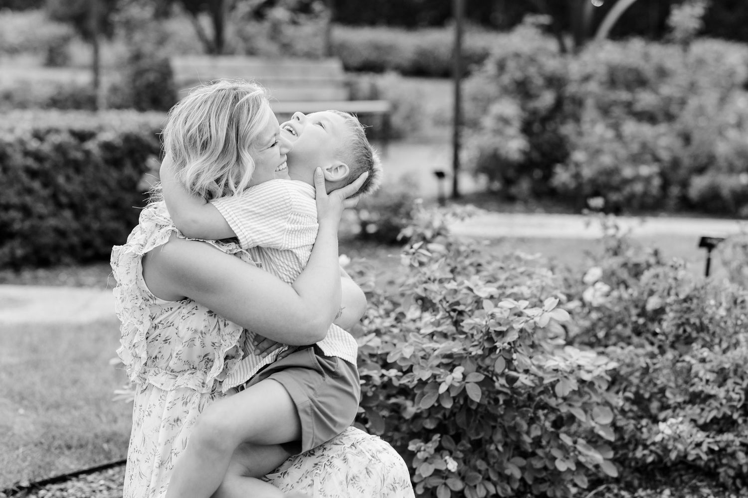 Black and white photo of a mother lovingly kissing her young child in a garden setting.