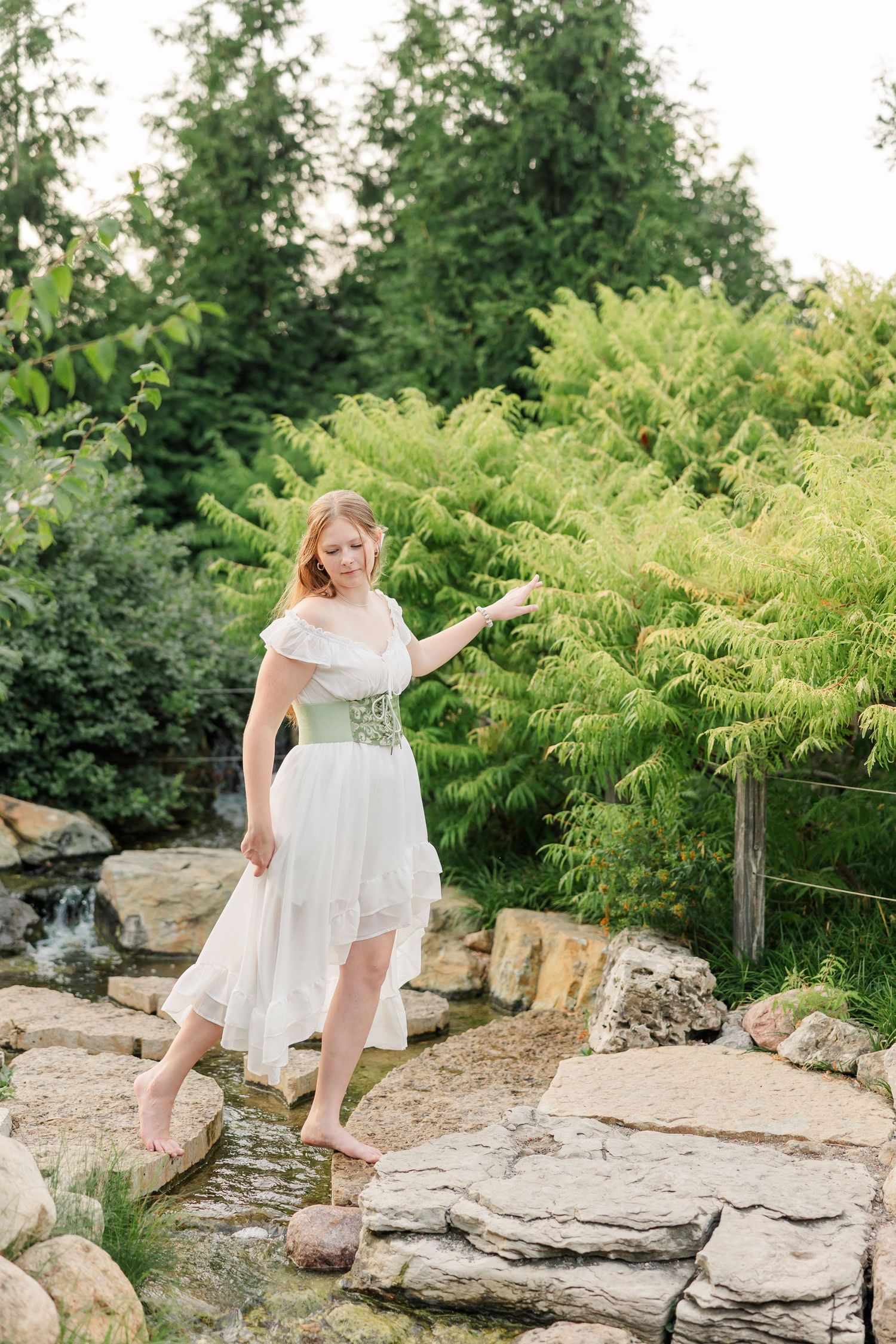 Samantha poses barefoot on garden rocks in a white dress and green corset during her Cantigny Park senior session.