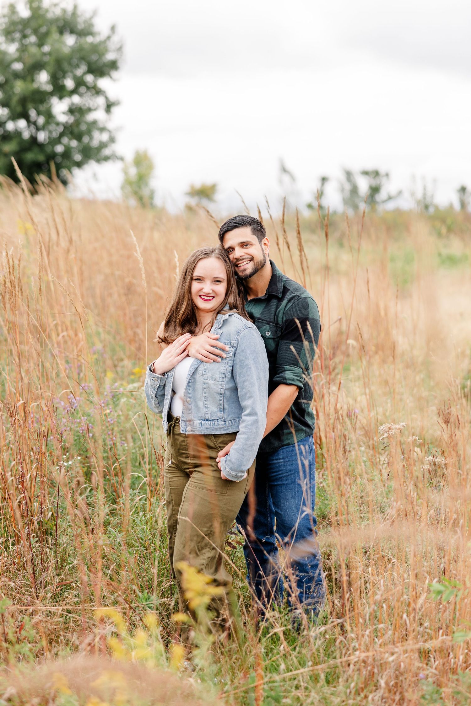 Smiling couple in denim and plaid embracing in a field of tall golden grass during an autumn engagement photo shoot.