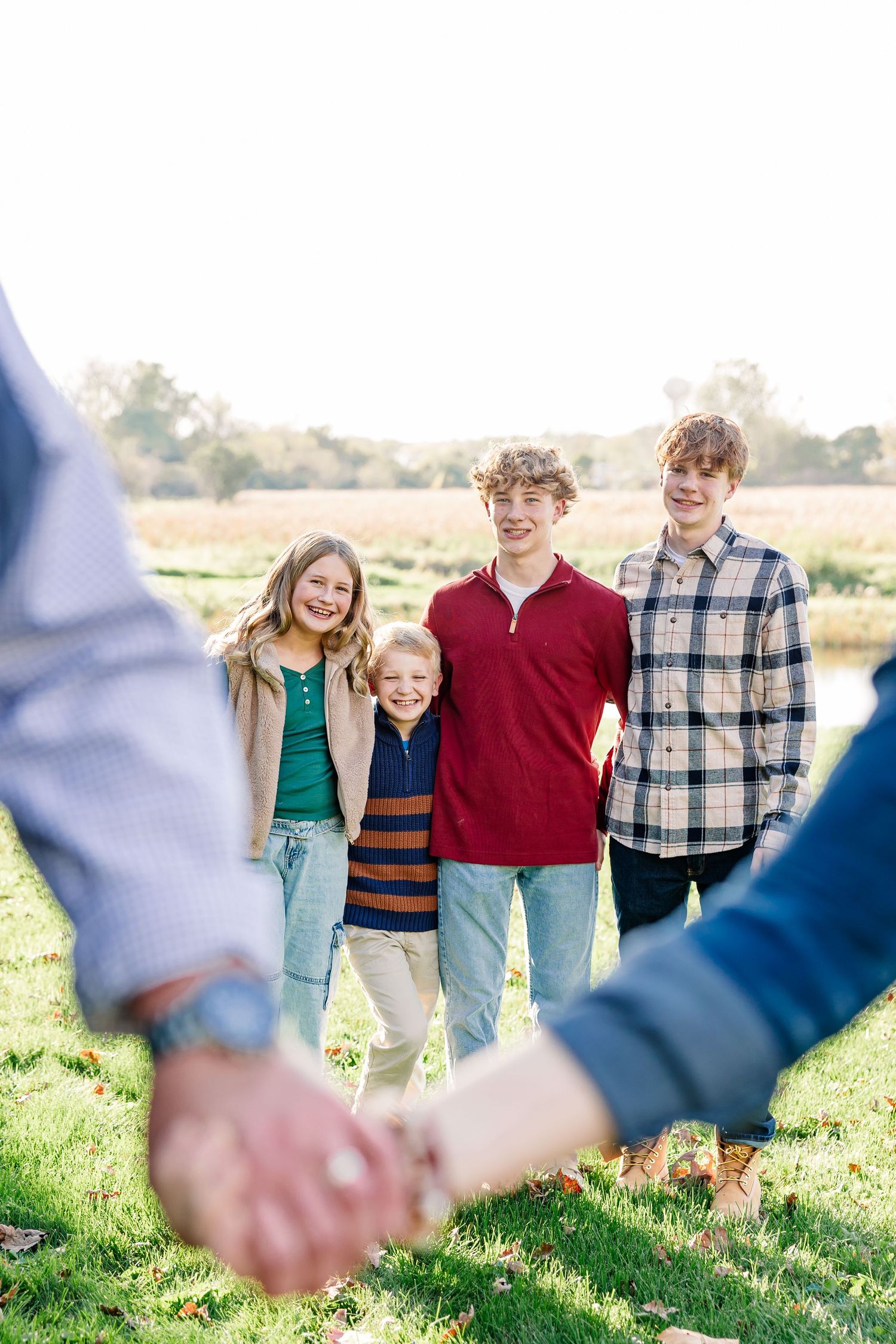 Four smiling siblings stand close together on a grassy field in autumn while two parents' clasped hands appear in the foreground.
