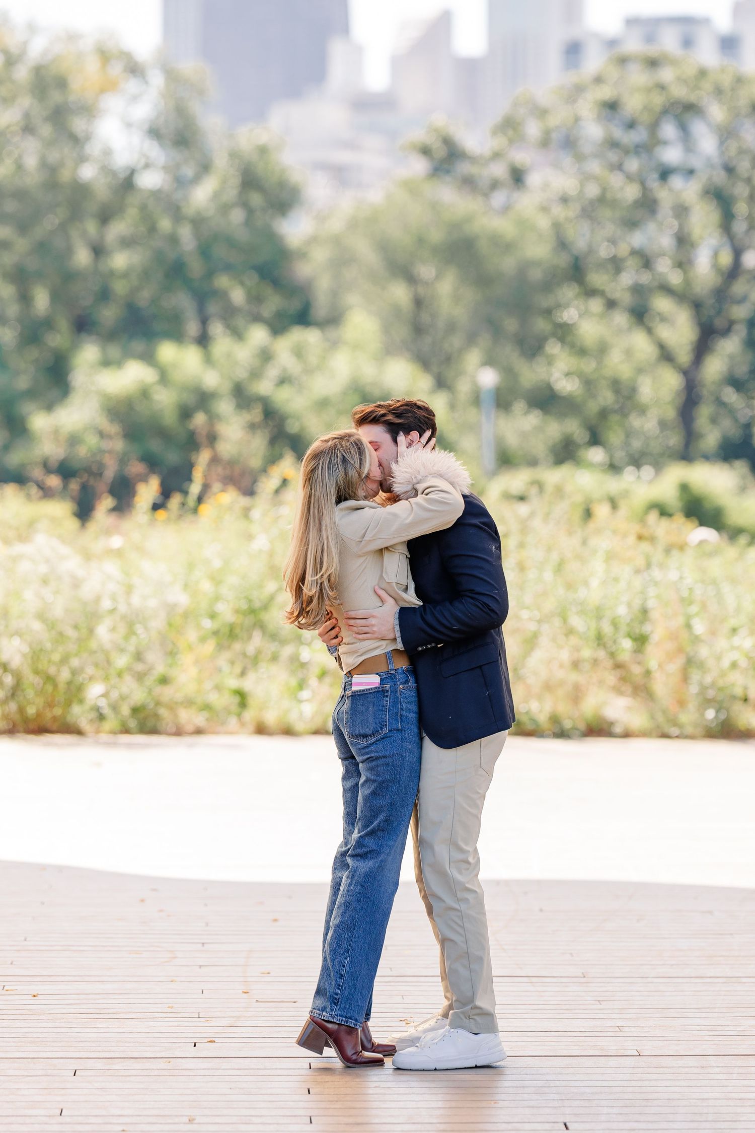 Couple sharing a romantic kiss on an outdoor deck surrounded by lush greenery and a city skyline backdrop.