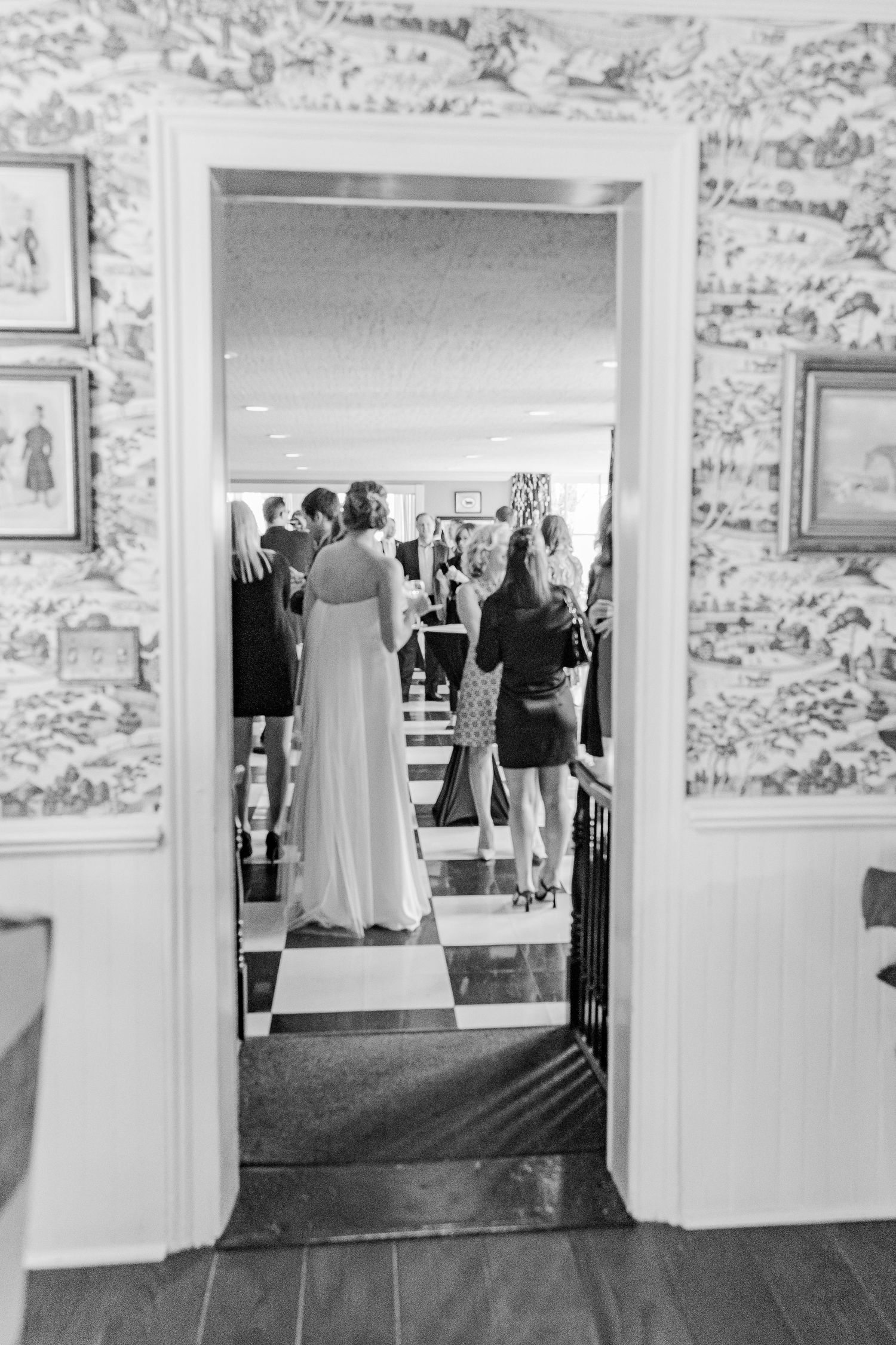 Black and white photo of a wedding gathering viewed through a doorway with floral wallpaper.