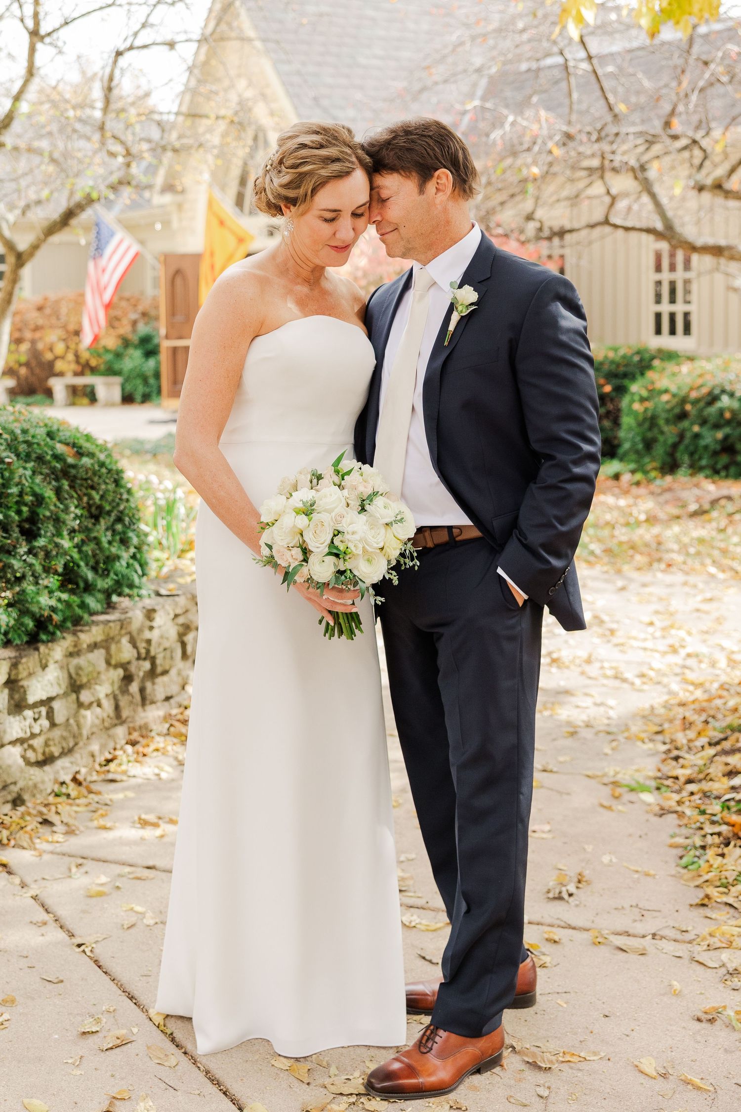 A romantic autumn wedding photo with couple standing on stone path surrounded by fall foliage.