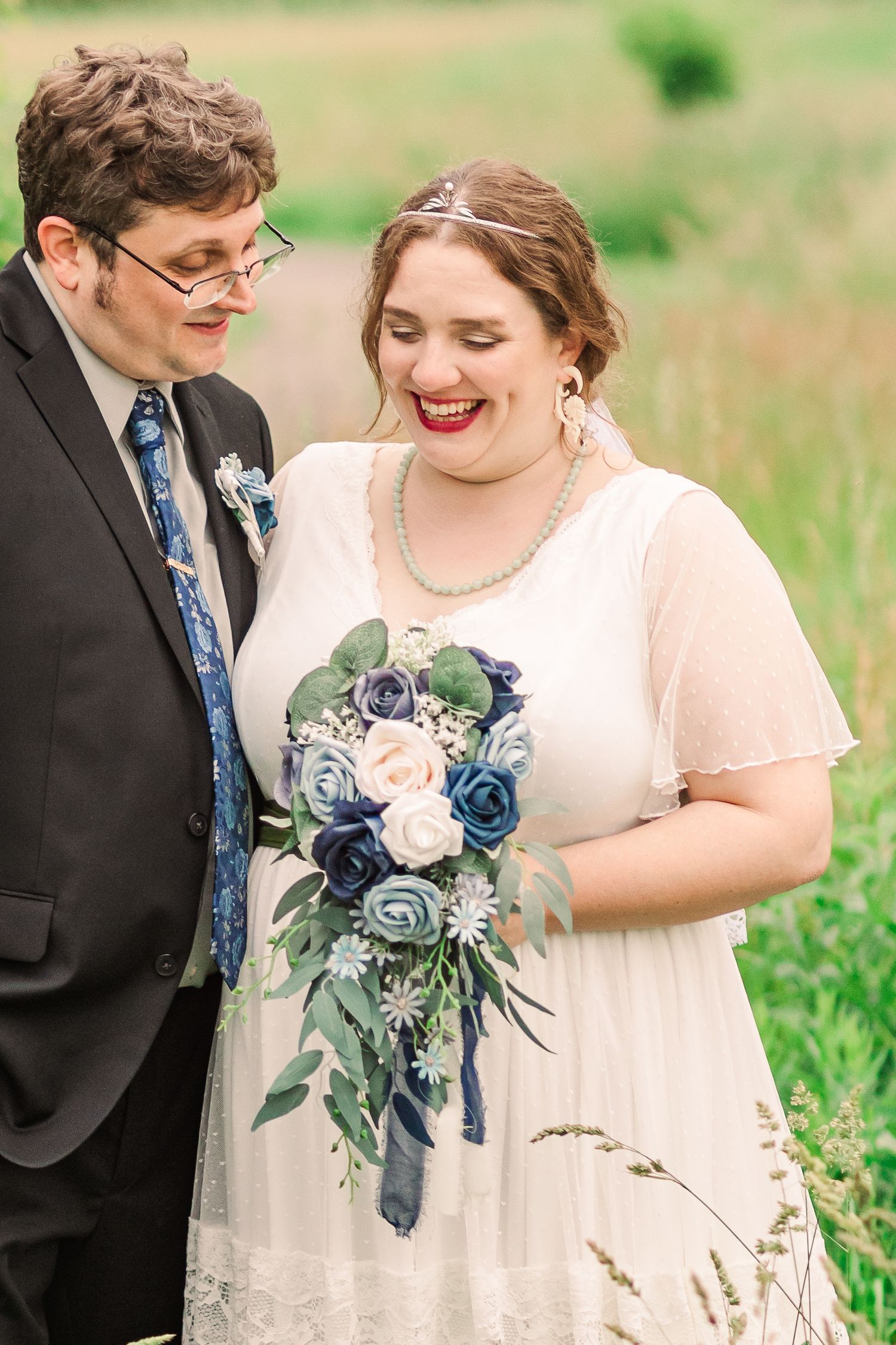 A wedding couple shares a joyful moment outdoors with the bride holding a blue and blush rose bouquet.