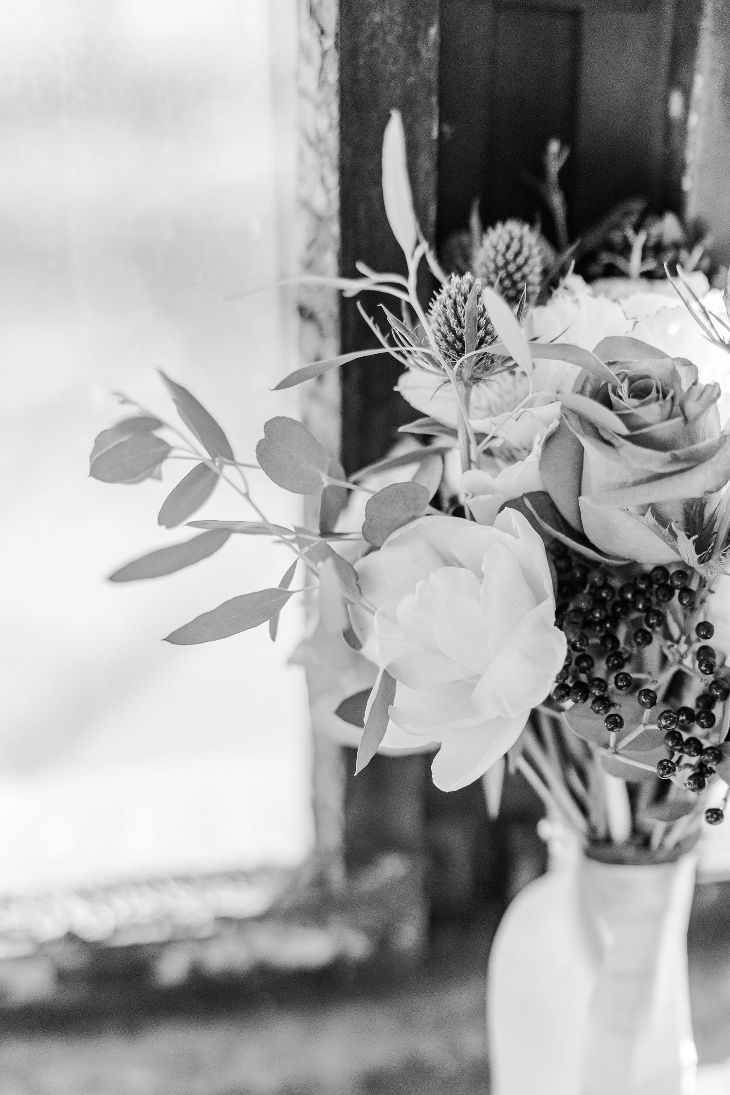 Black and white photograph of elegant roses and thistle flowers arranged in a vase by a window.