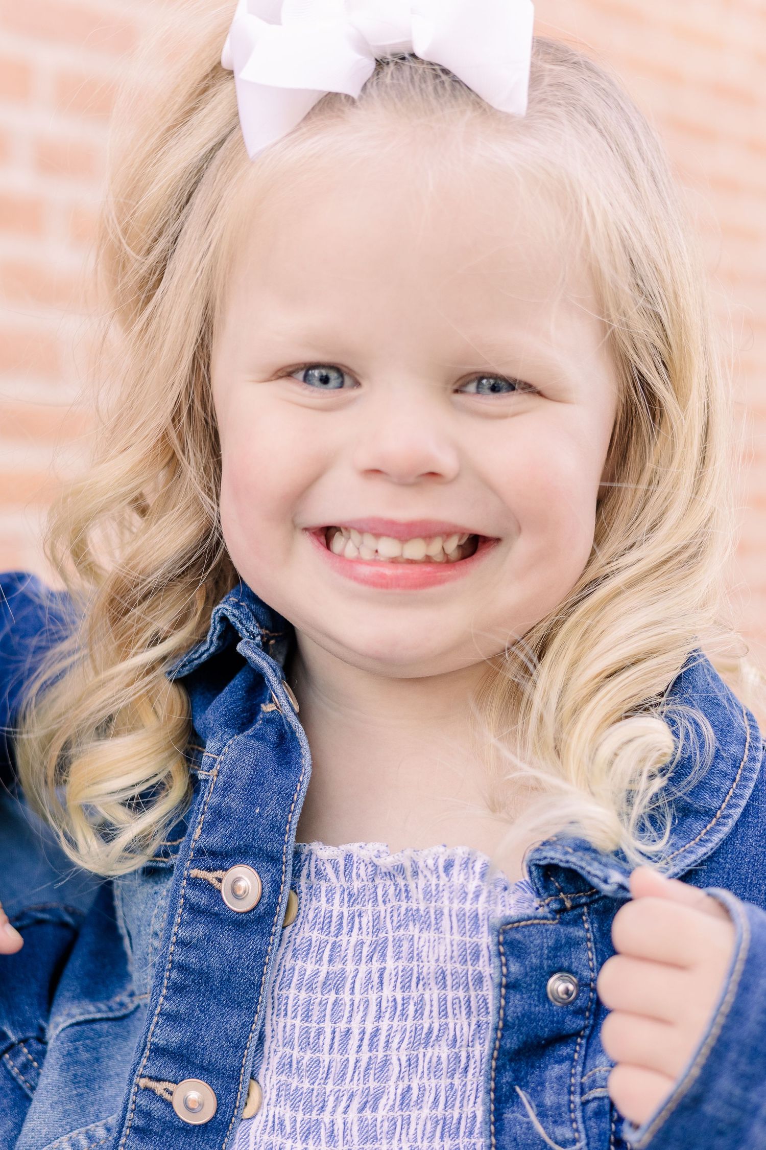 Young child wearing denim jacket and white bow smiling at camera with blonde curly hair against brick wall background.