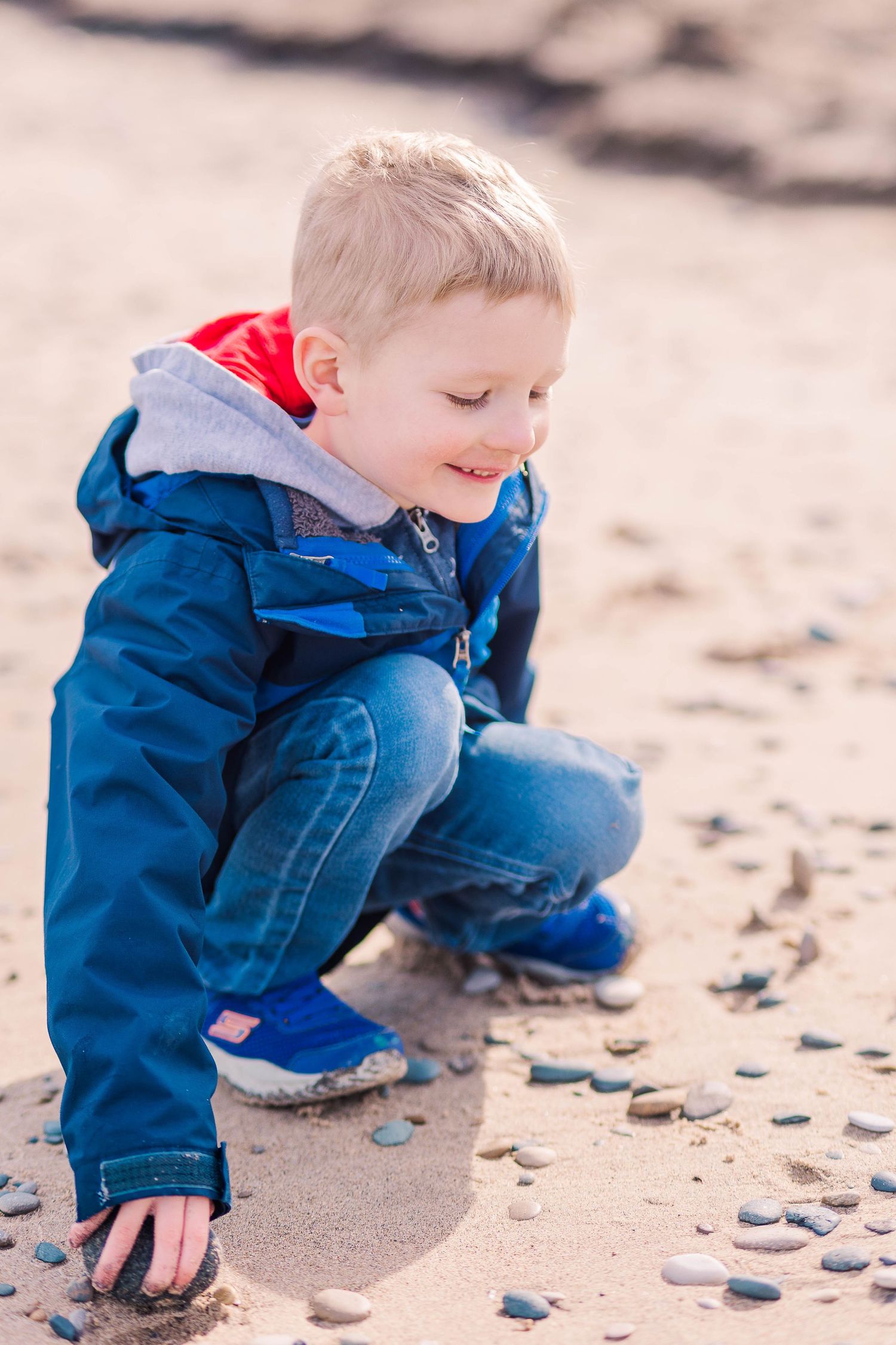 Active child in blue winter coat crouches down to explore pebbles on sandy beach.