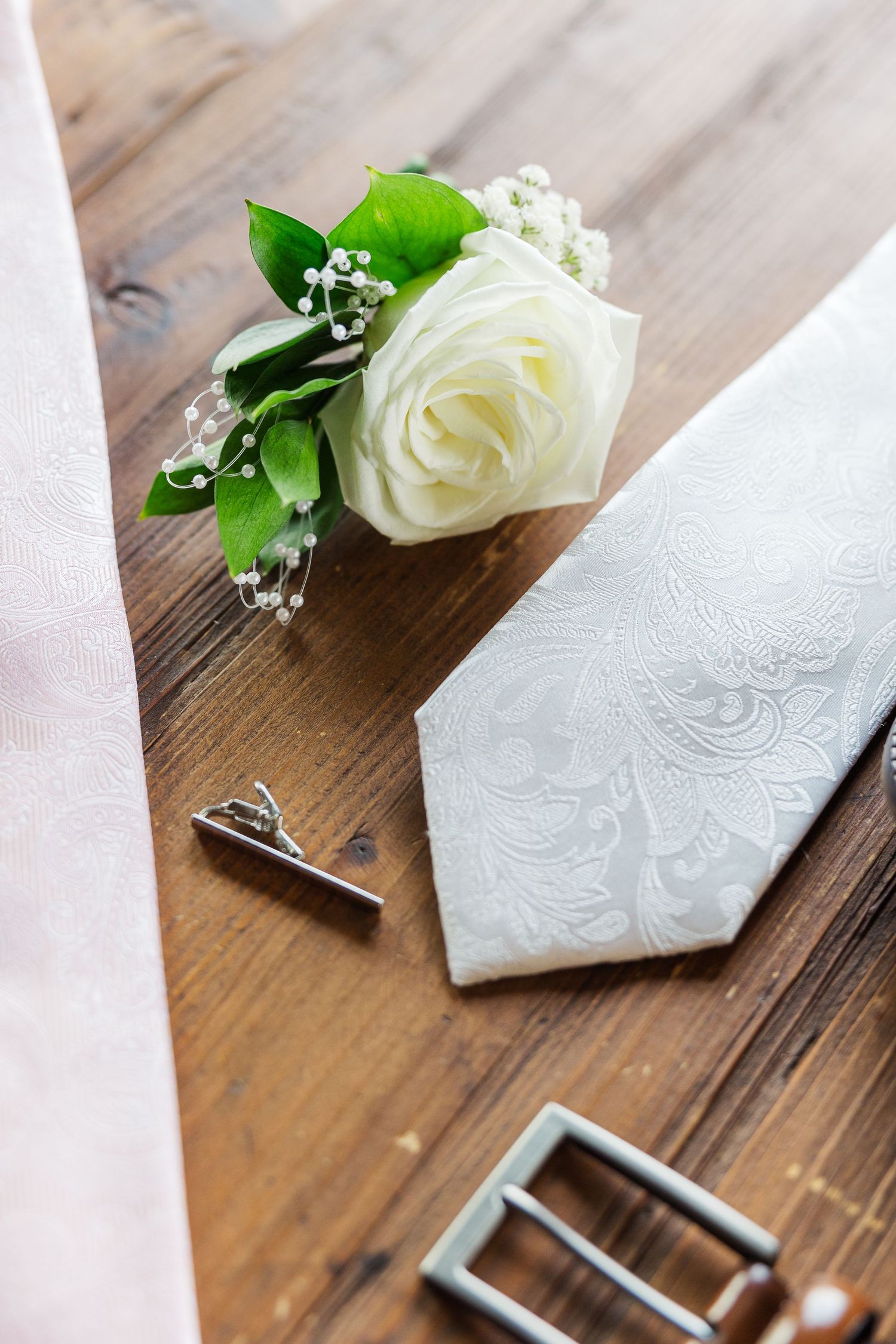 White rose boutonniere with baby's breath next to a white tie and silver belt buckle on wooden surface.