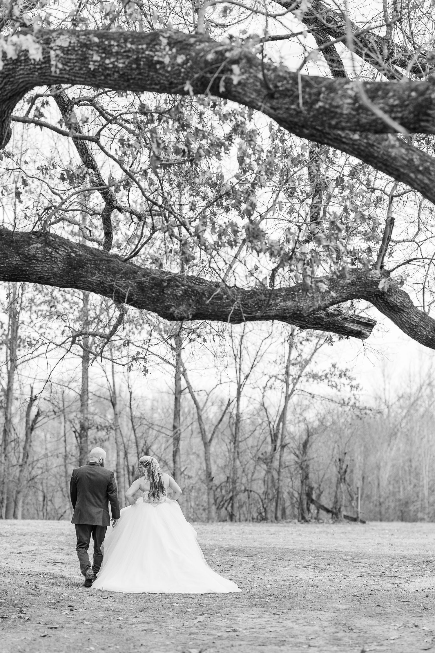 Black and white wedding photograph showing couple walking under arching tree branches.