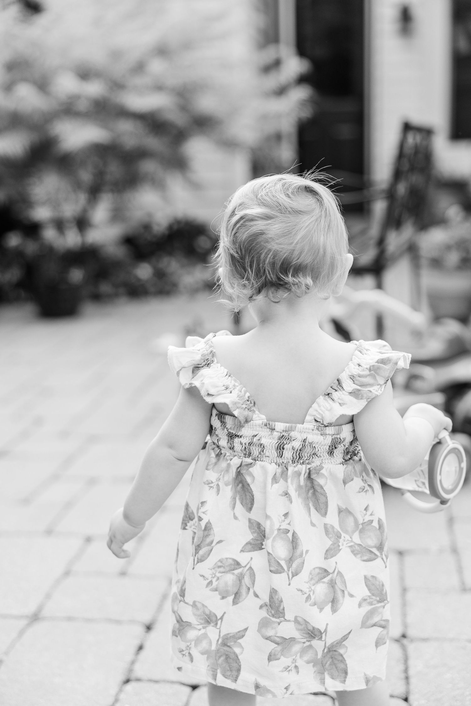 A toddler in a floral dress walking away on a stone patio.