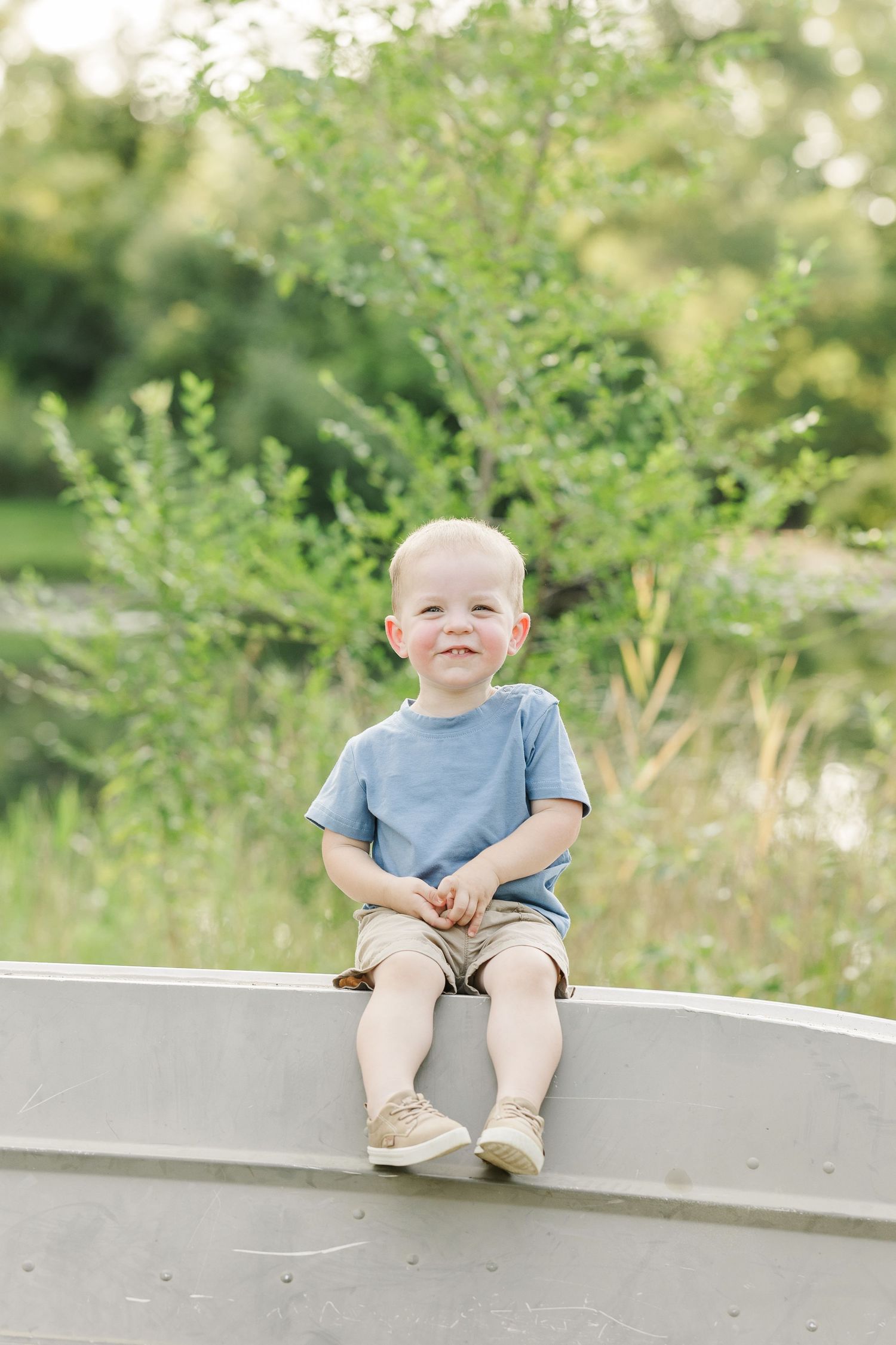 A child in a light blue t-shirt sits on a curved concrete structure amidst green foliage on a sunny summer day.