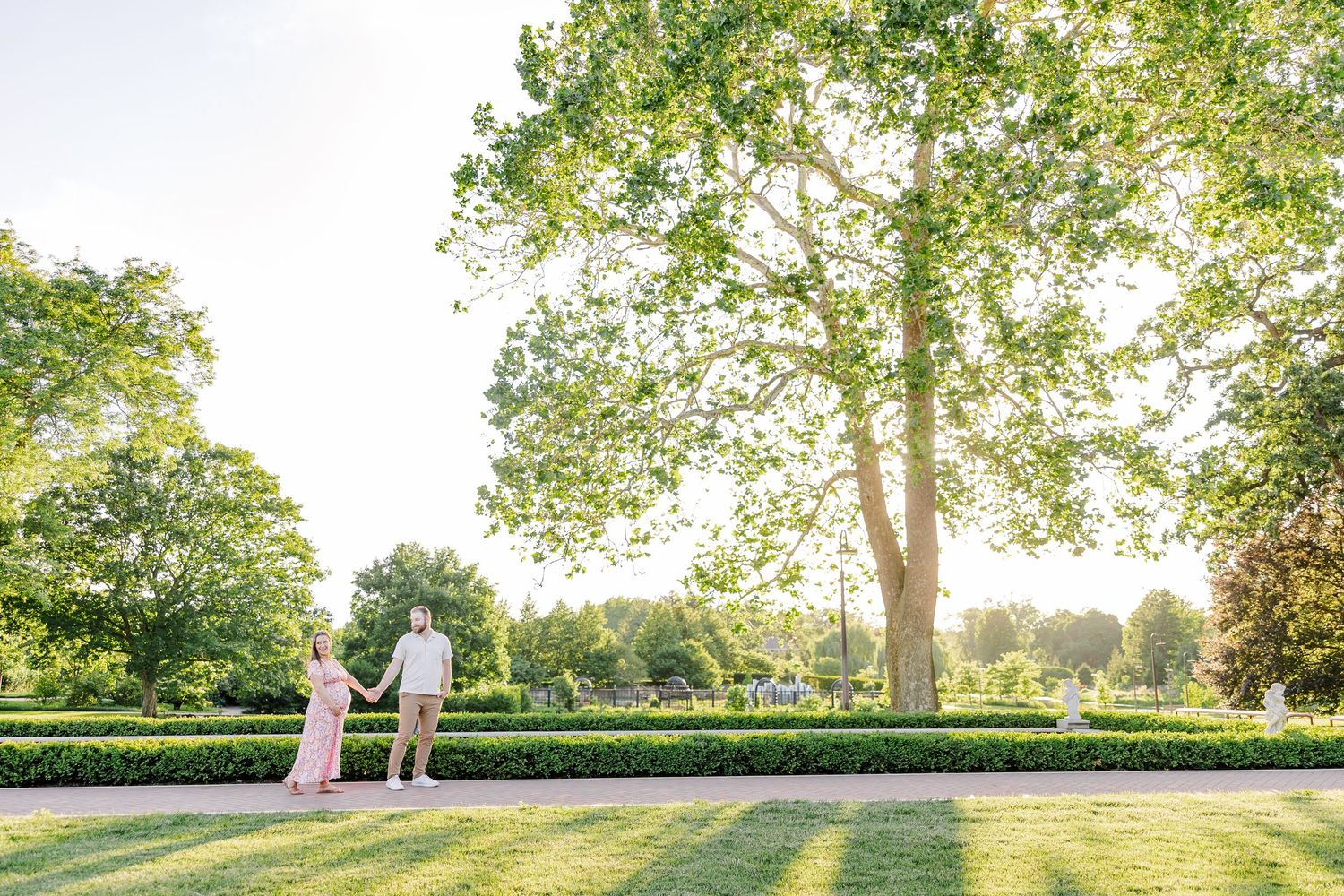 Wide Cantigny maternity photo of parents-to-be walking near garden benches in Wheaton Illinois summer light