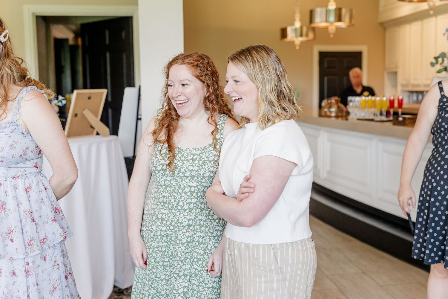 Two young women smiling and laughing together at an indoor celebration event in a bright, elegant venue.