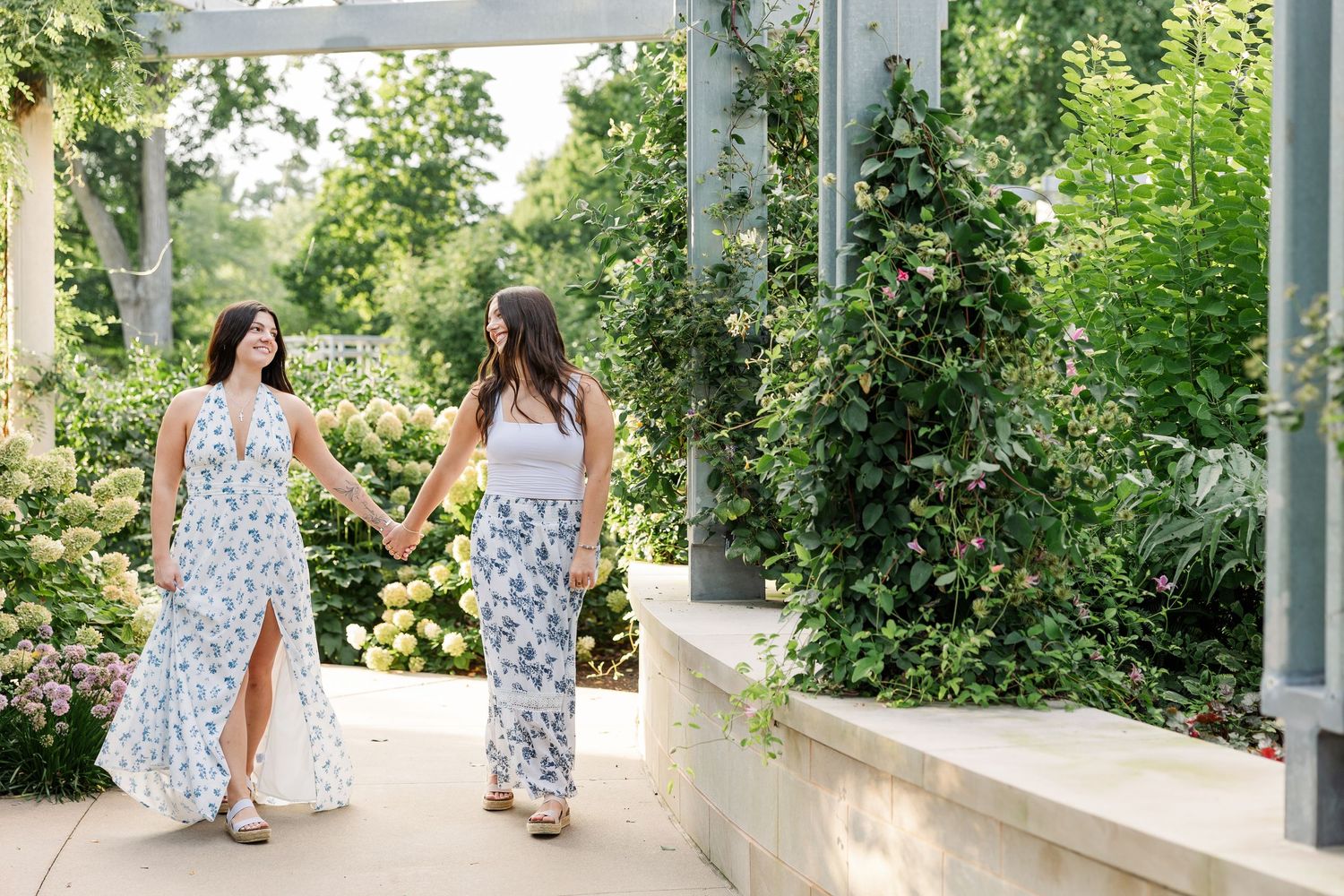 Two women in floral maxi dresses walk hand in hand through garden pergola.