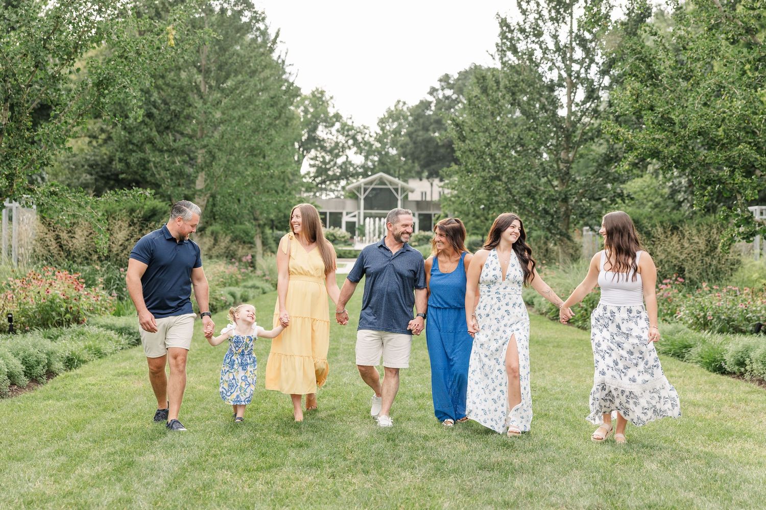 Family of six walking hand-in-hand through a lush garden, dressed in coordinated summer outfits, smiling outdoors.