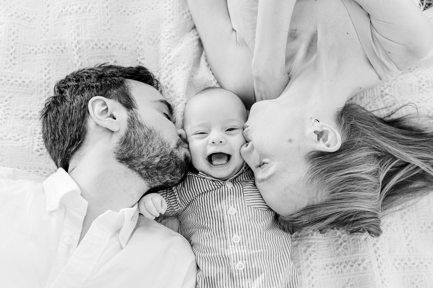 Black and white photo of parents kissing laughing newborn baby lying on white blanket.