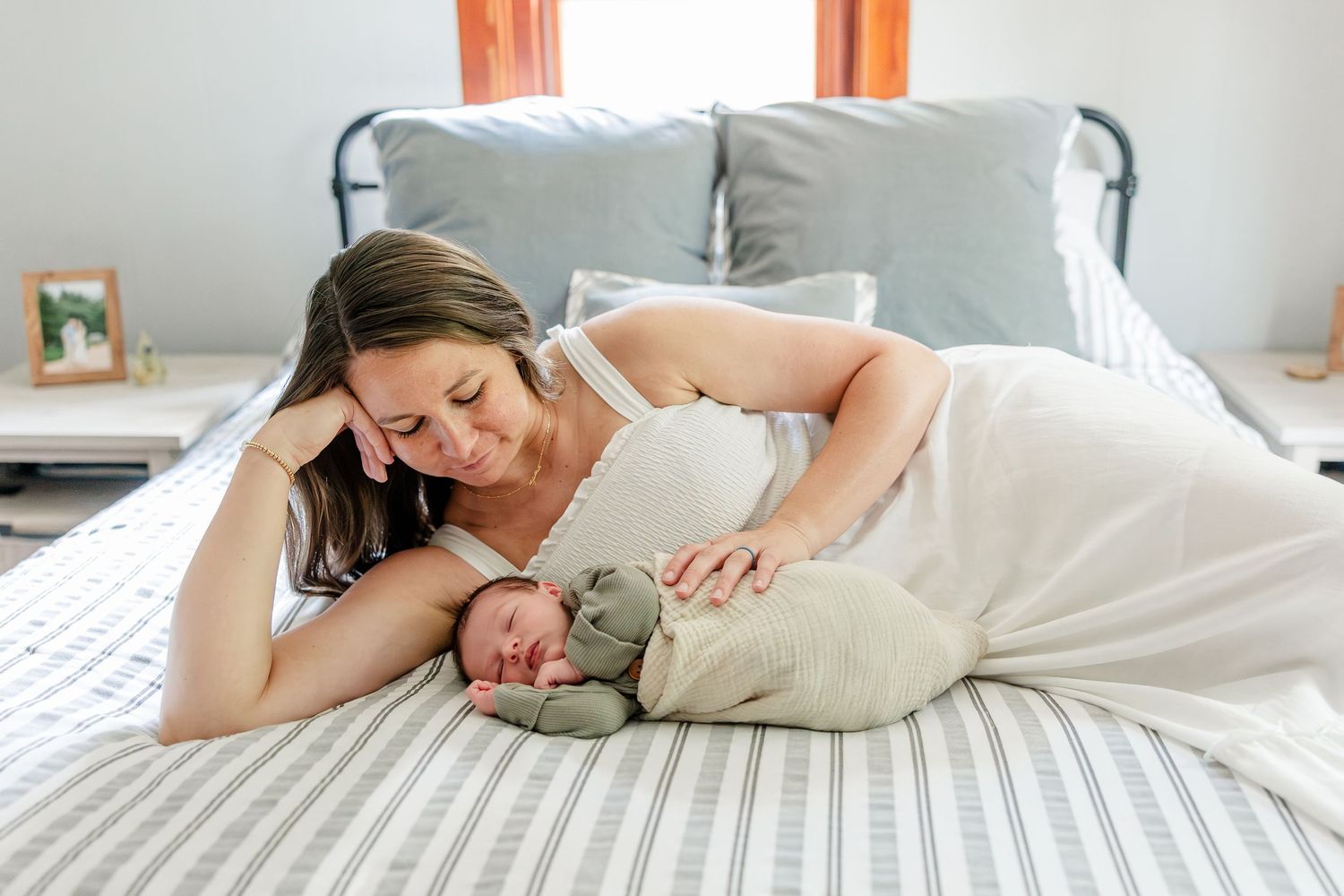 A mother lying on a bed smiles lovingly at her swaddled newborn baby during an in-home newborn photography session.