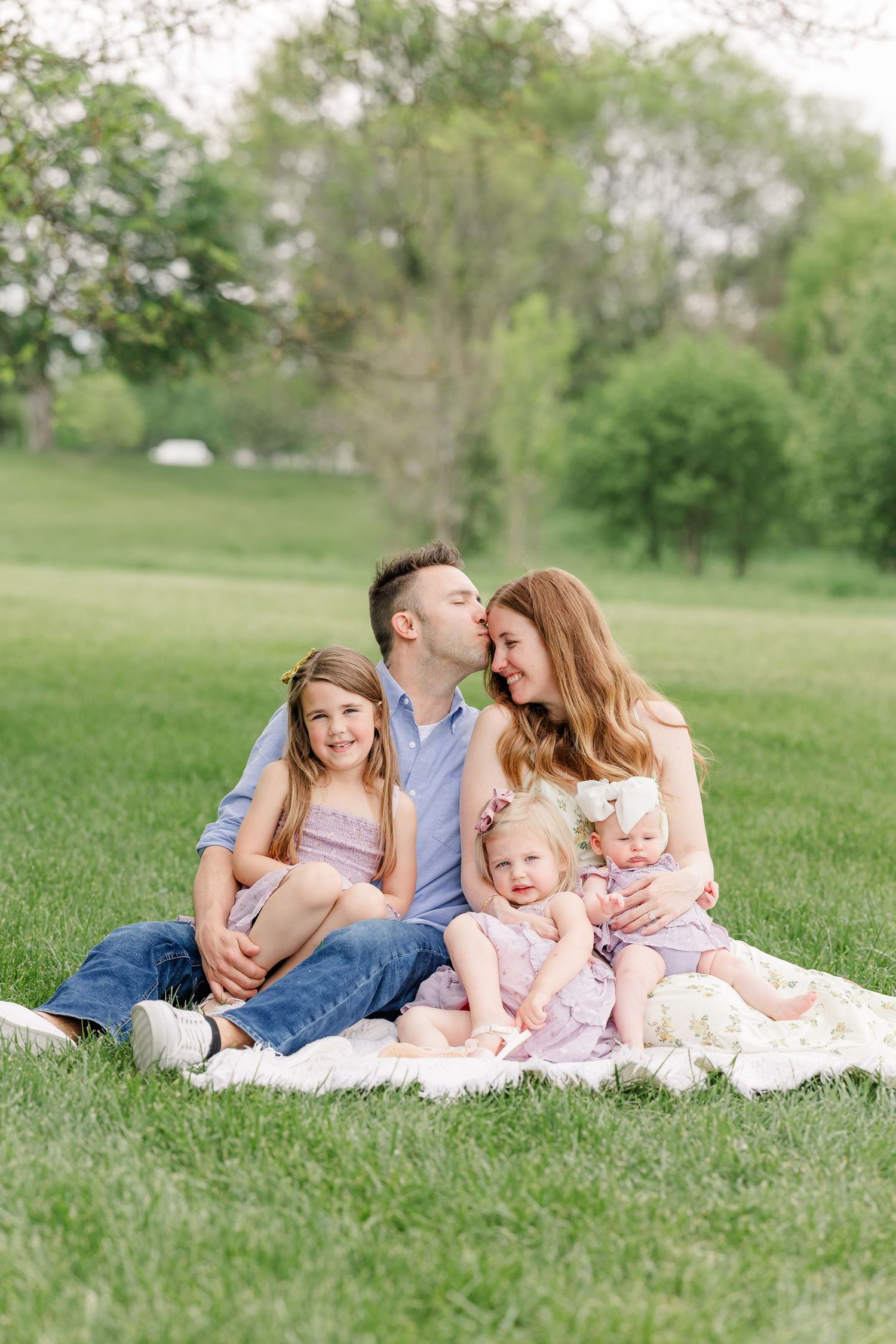 Family of five sitting on a white blanket in a green park with father kissing mother on the cheek.