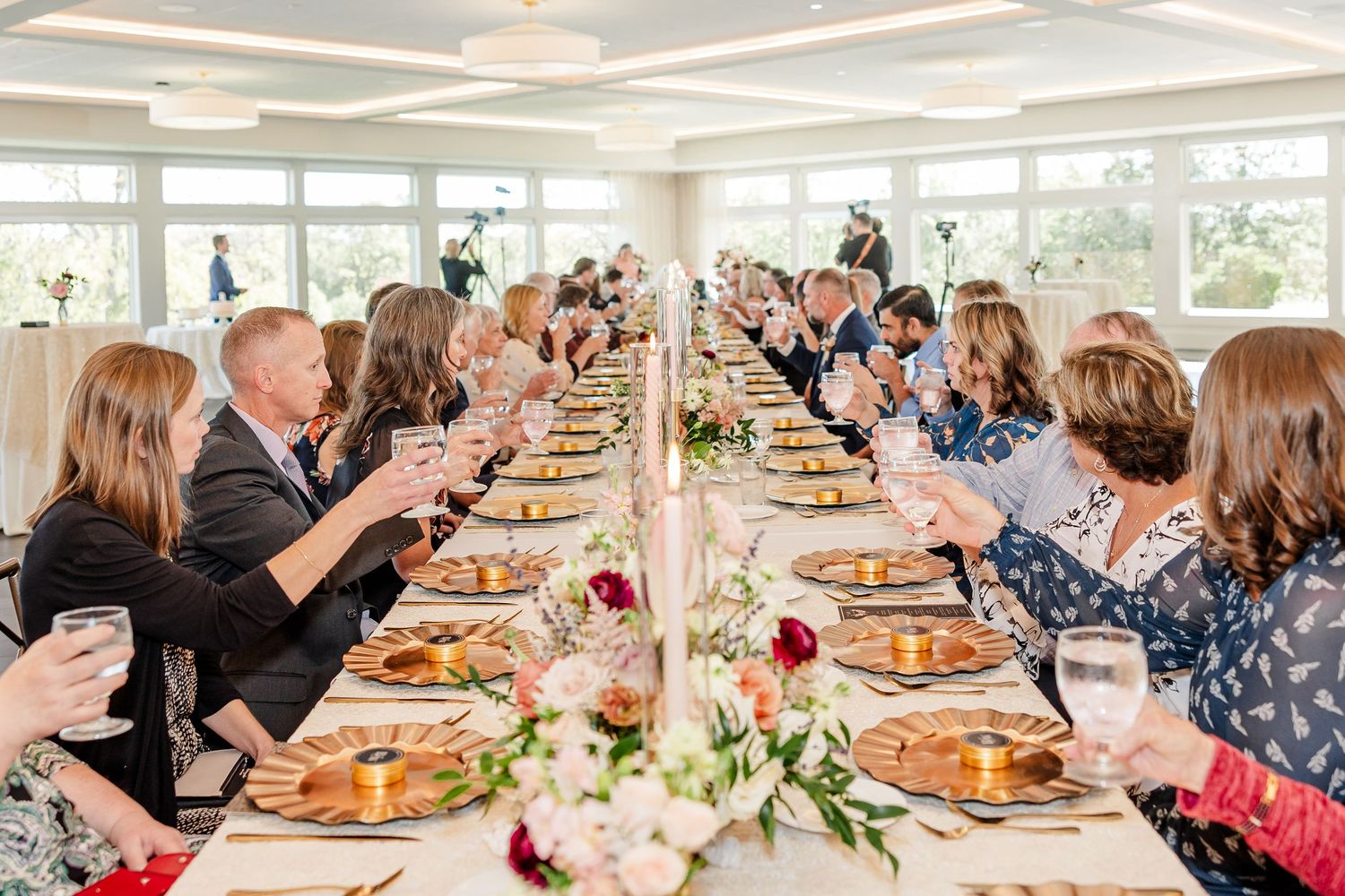 Wedding guests seated at a long reception table with floral centerpieces and candles raise glasses in a toast near large windows.