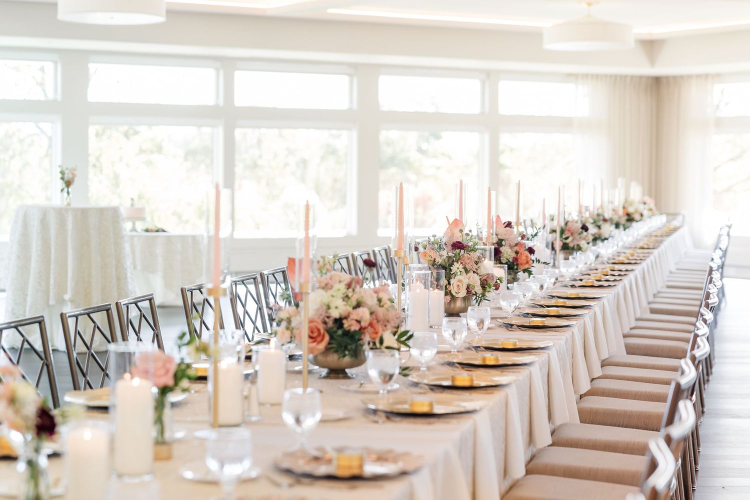 Long wedding reception table with white linens, candles, floral centerpieces, and place settings in a bright room with large windows.