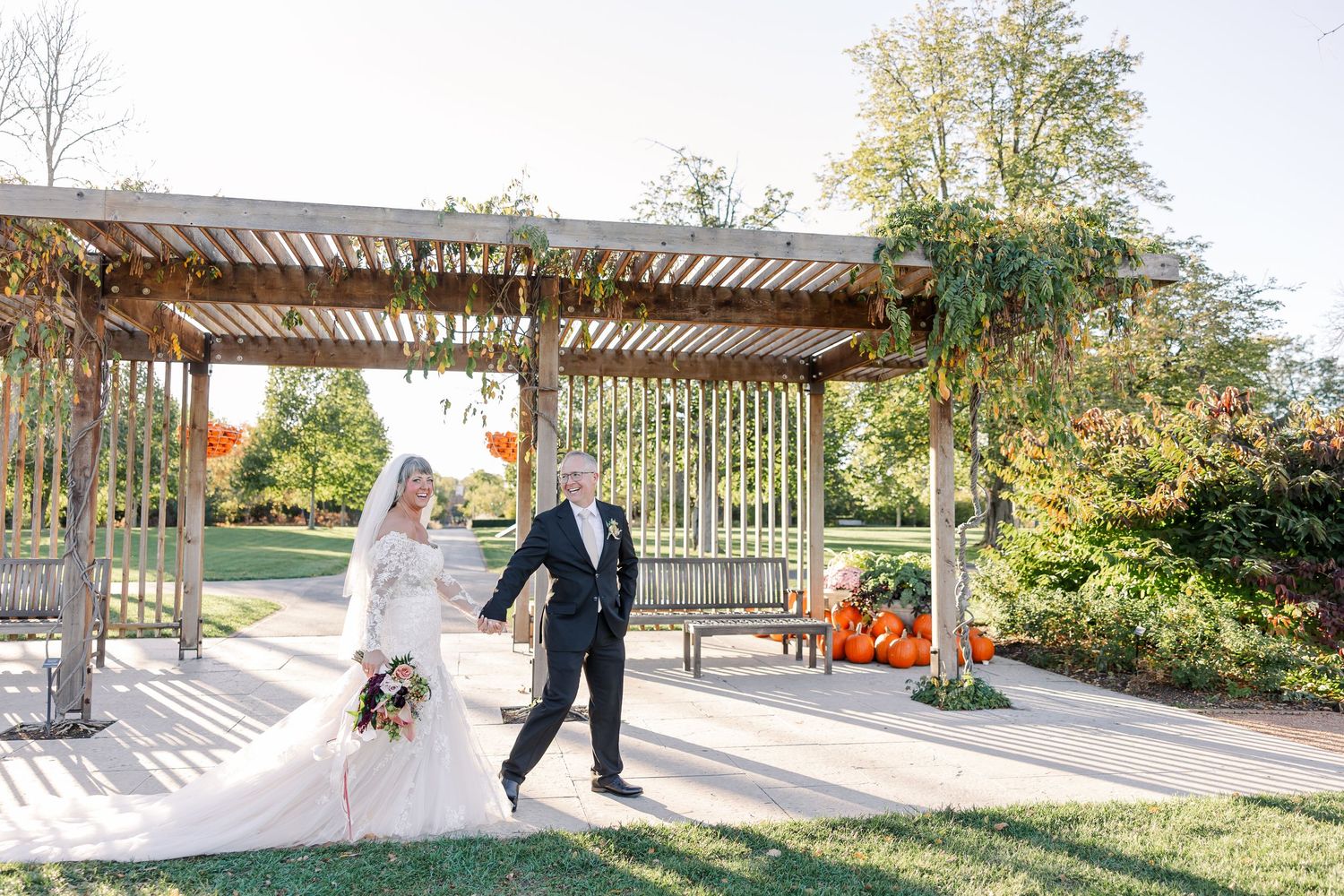 Bride in a lace wedding gown holding a bouquet walks hand in hand with a groom in a dark suit past a wooden pergola on a sunny autumn day.