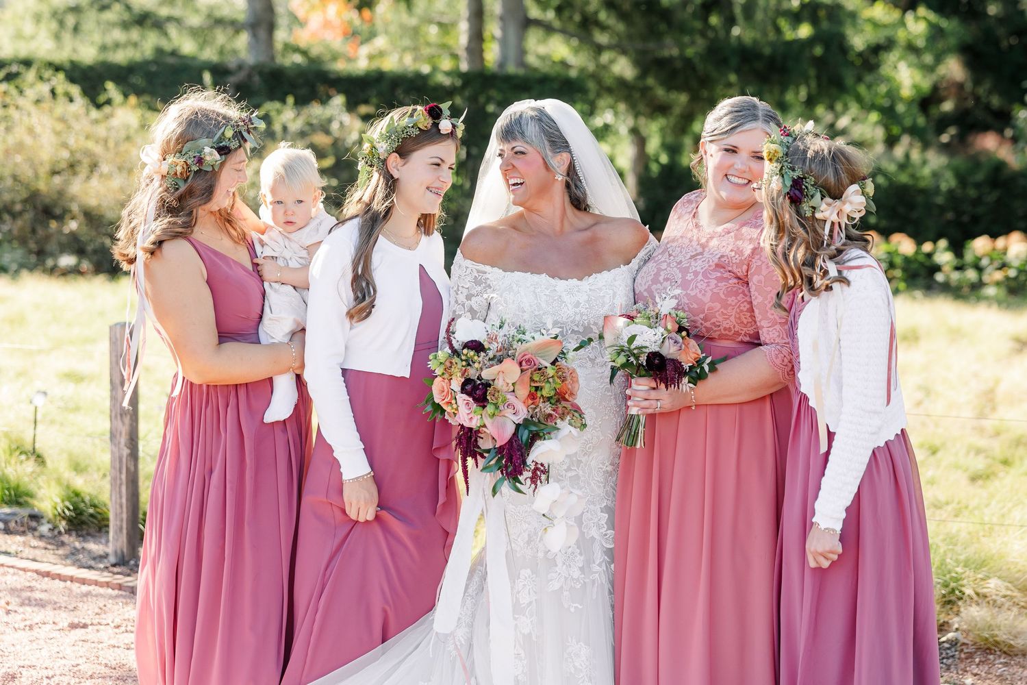 Bride in a white lace gown and veil laughs with bridesmaids in dusty rose dresses and floral crowns, holding bouquets outdoors.