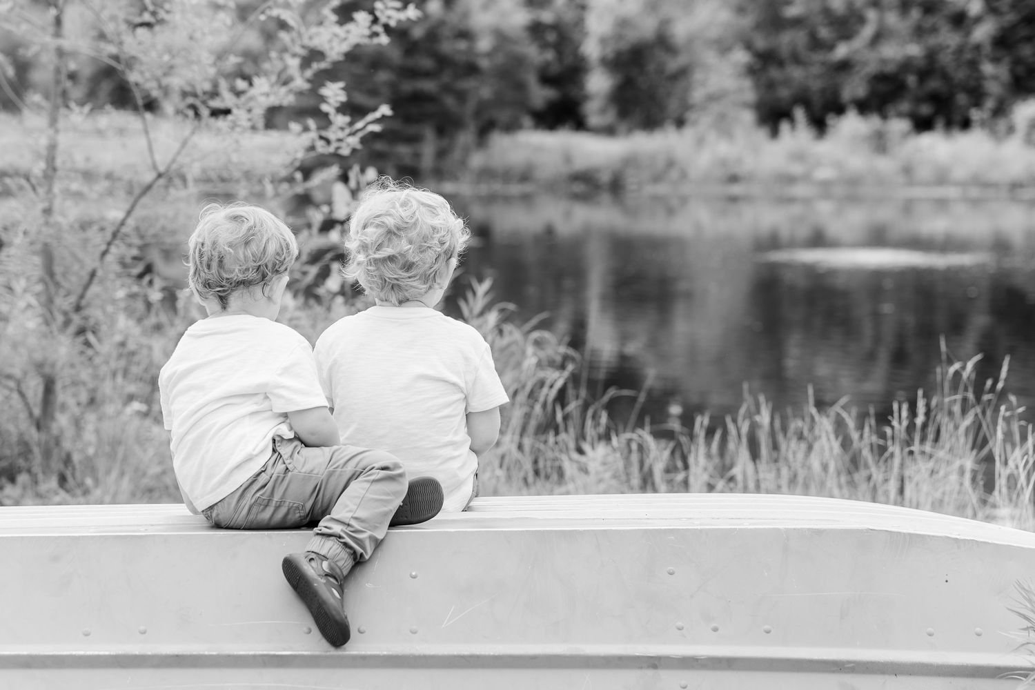 Two young boys sit together on a boat edge, gazing at a serene lake surrounded by lush greenery.