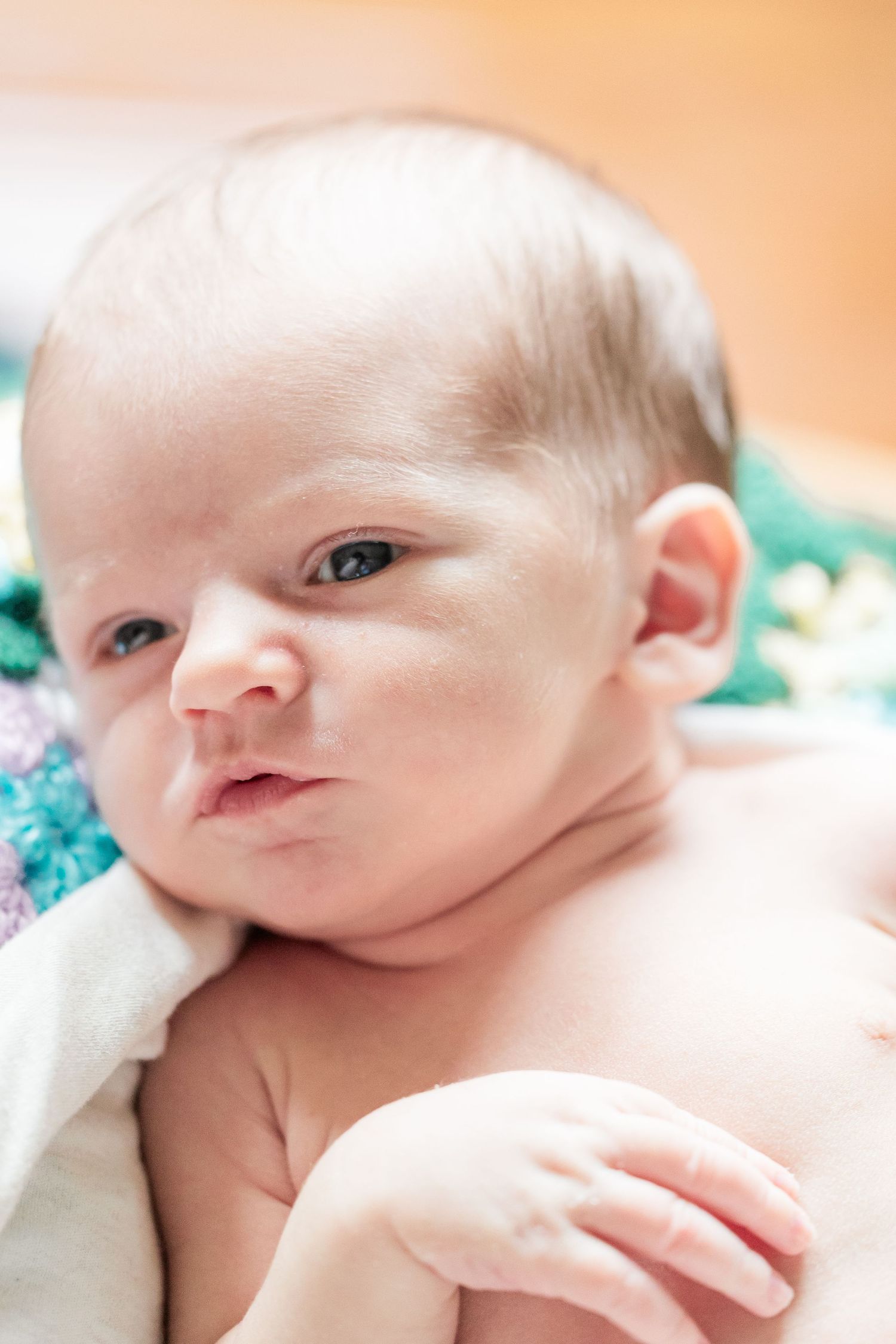 Close-up portrait of alert newborn baby with dark hair lying on colorful blanket, eyes open.
