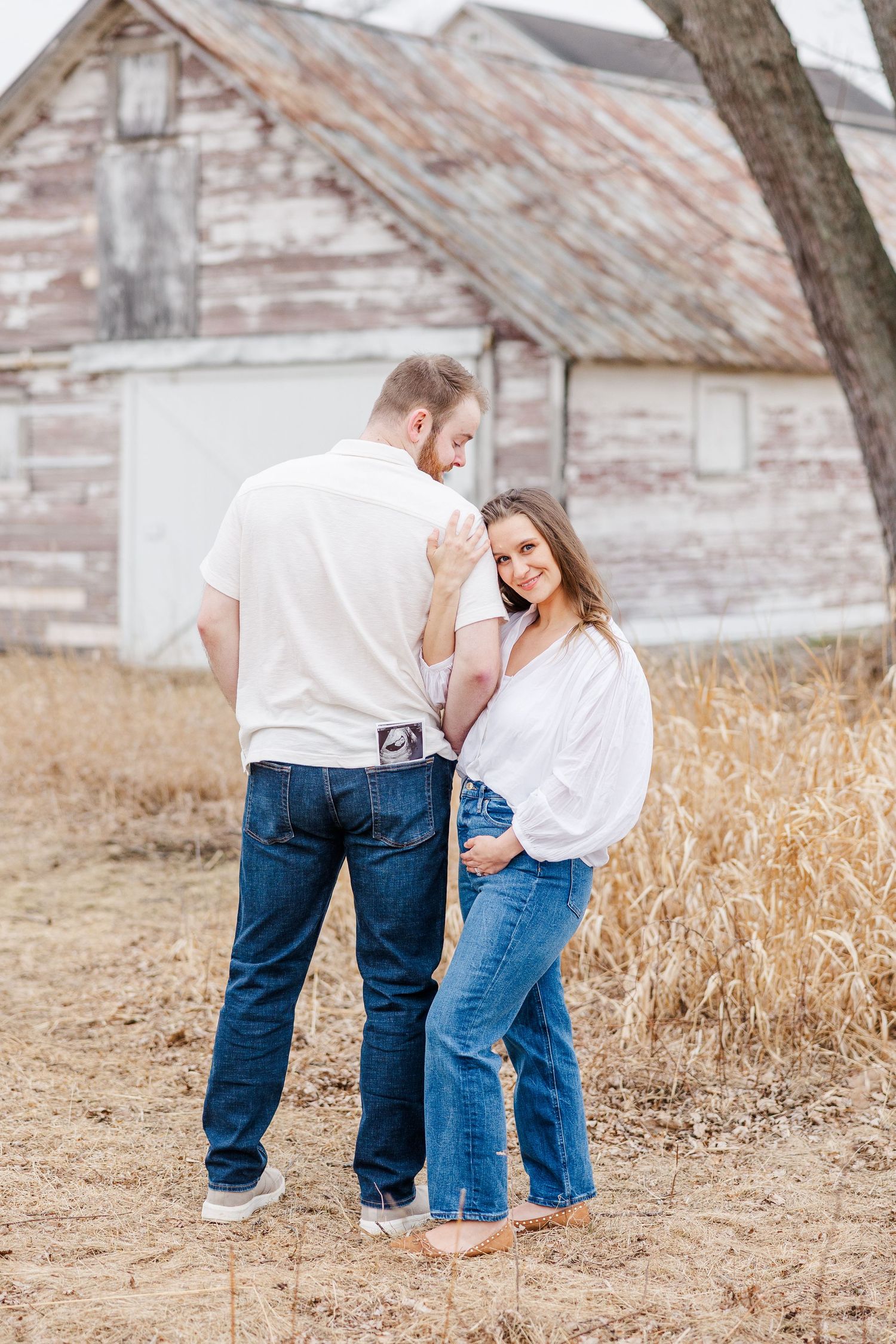 Couple posing in tall grass by rustic barn during pregnancy announcement photos in Cary, Illinois