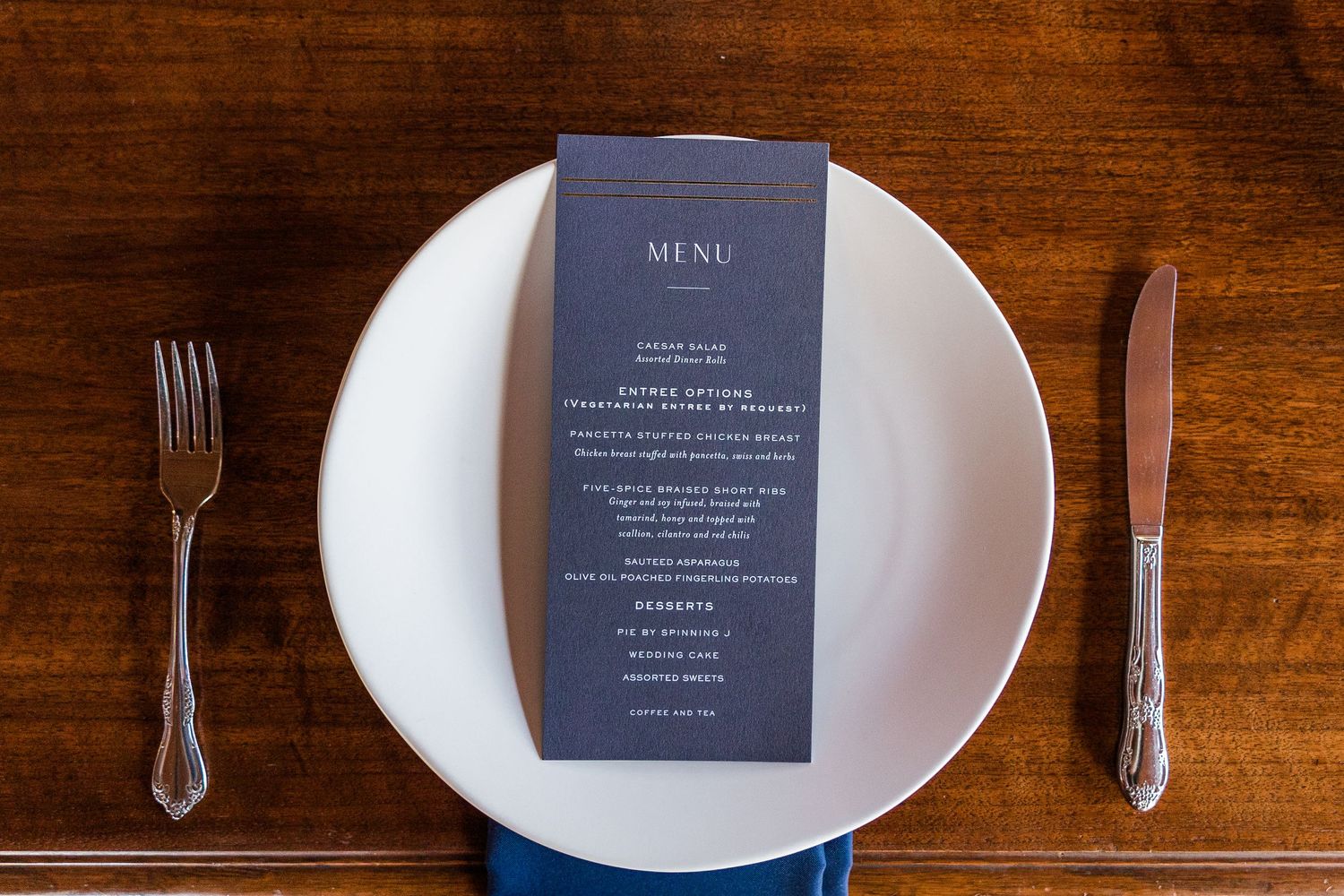 Elegant table setting with white plate, silverware and black menu card on wooden surface.