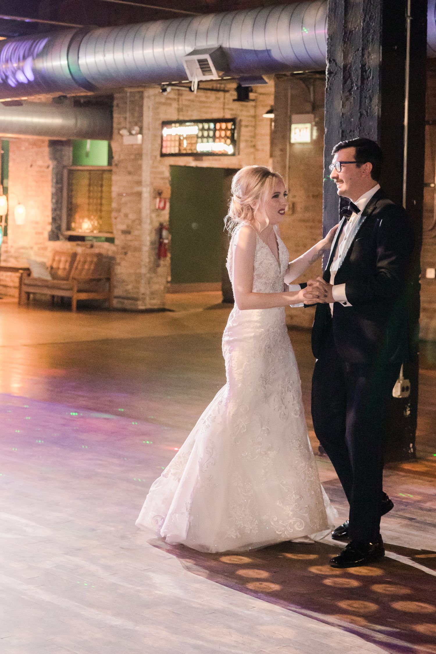 A couple sharing their first dance at a rustic industrial wedding venue with exposed brick walls and overhead pipes.