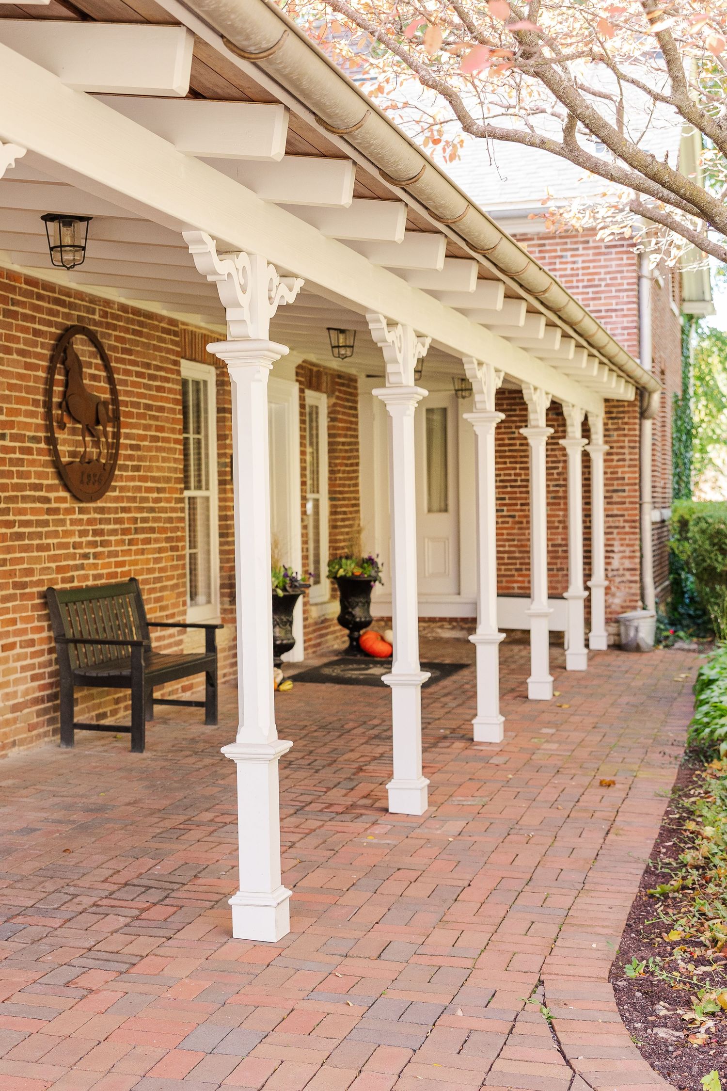 White columned porch with decorative details along brick building featuring benches and seasonal decorations.