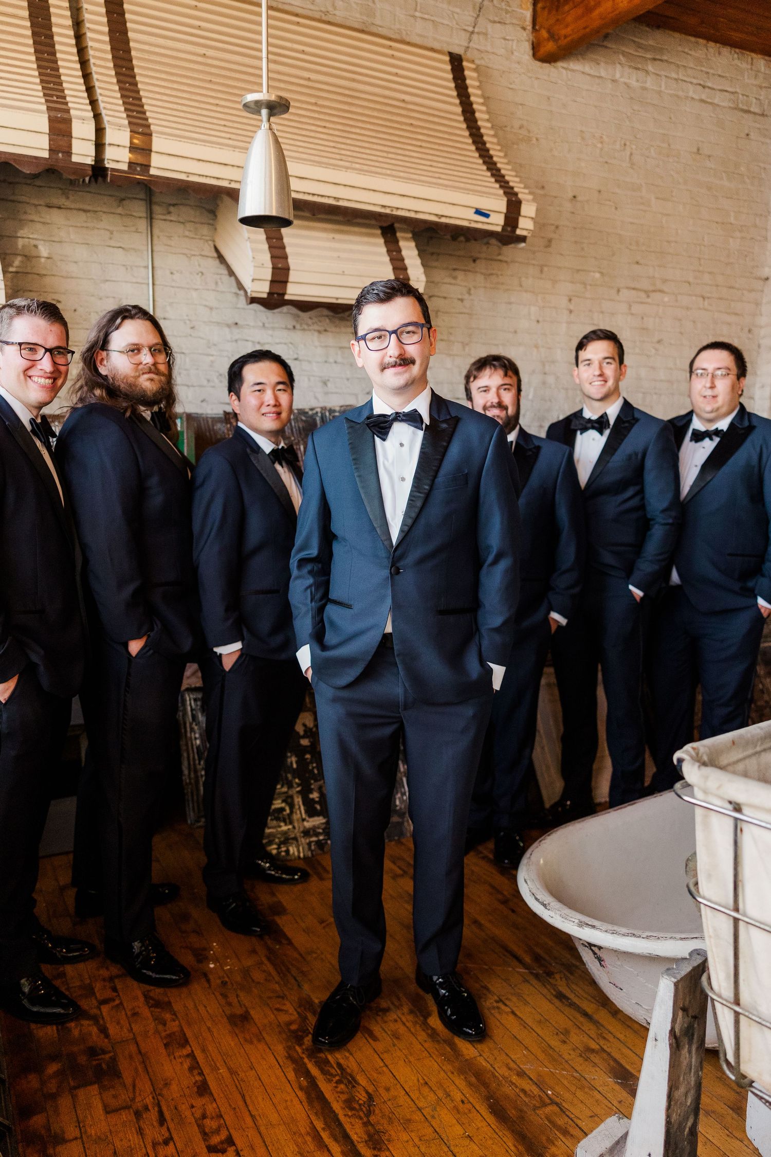 Groomsmen in formal attire pose together in a rustic room with wooden floors and exposed brick walls.