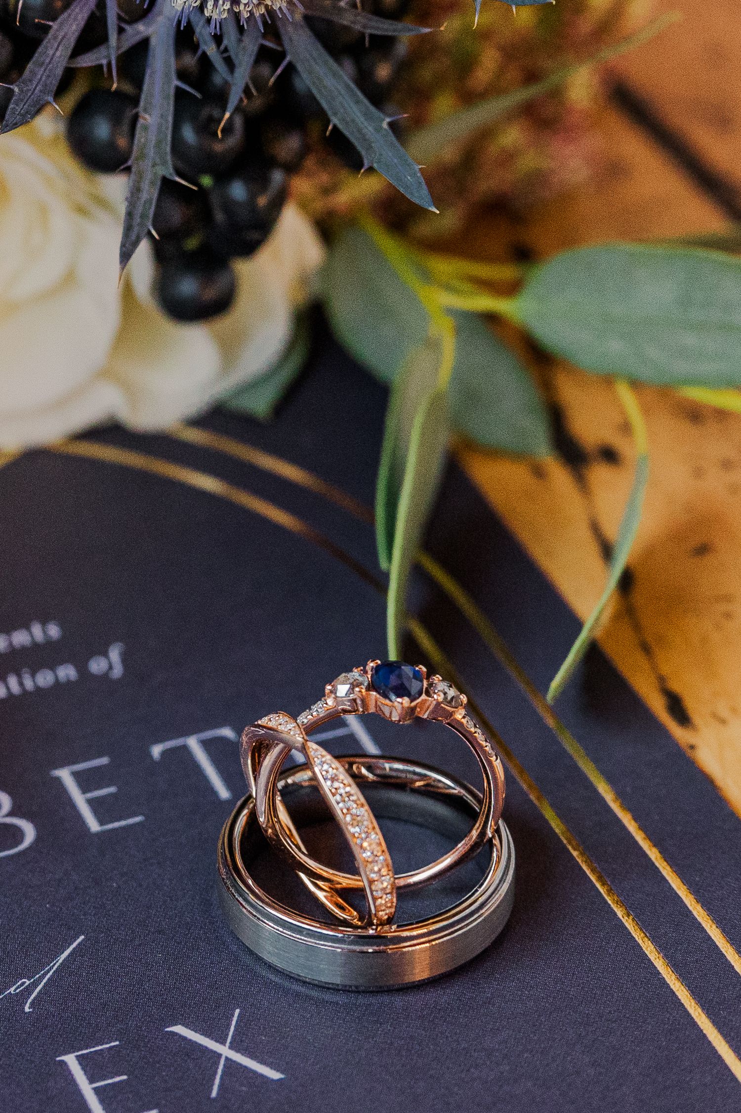 Diamond and sapphire wedding ring set arranged on dark invitation with floral elements.