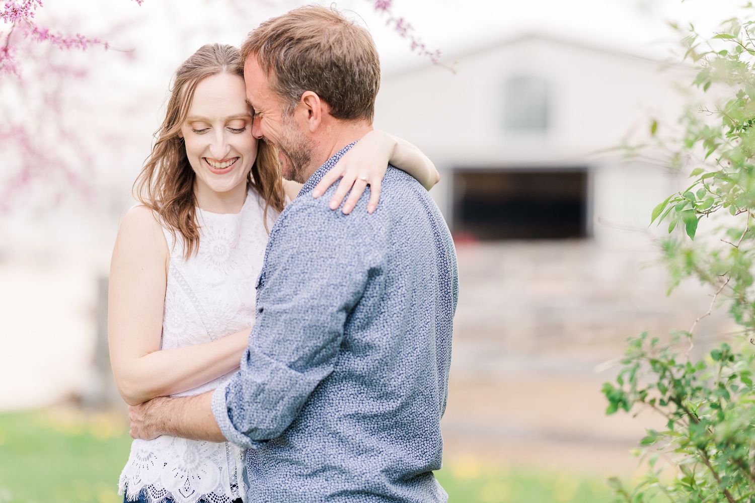Intimate couple moment captured near pink spring blossoms and white barn.
