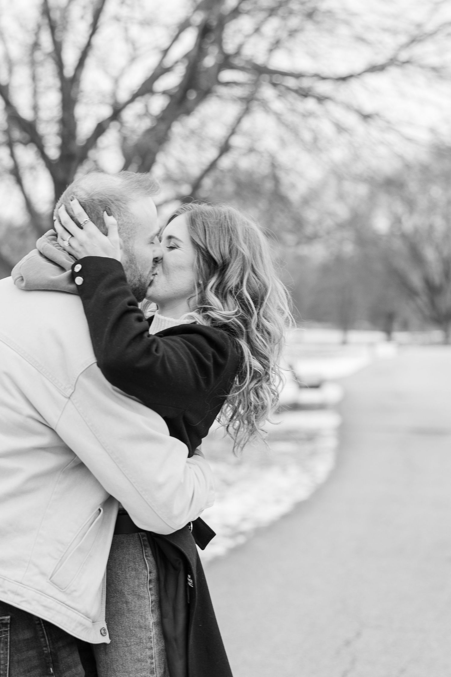 A romantic black and white photo of a couple embracing and kissing outdoors in winter with bare trees in background.