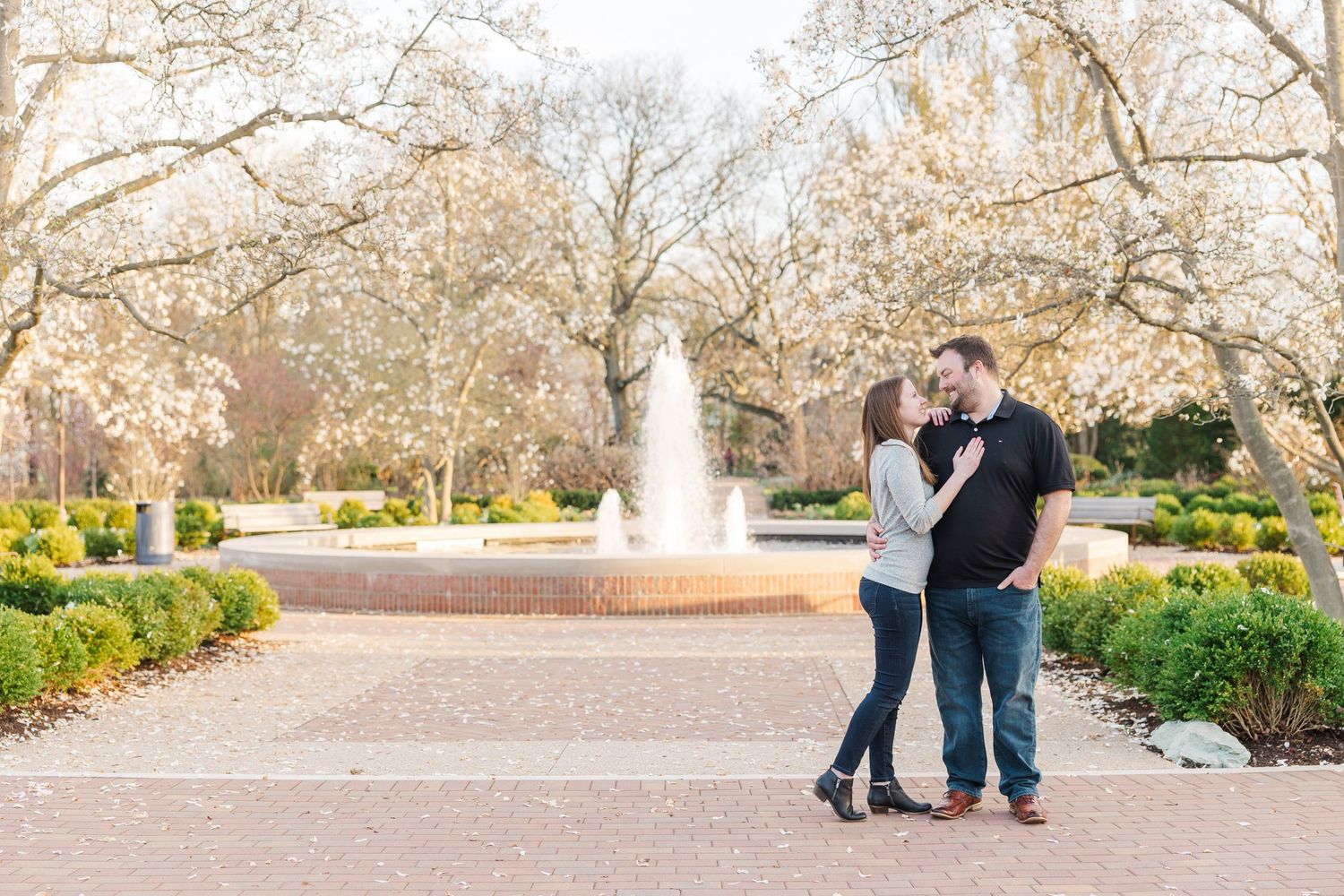 Romantic couple pose by fountain in garden with blooming trees.
